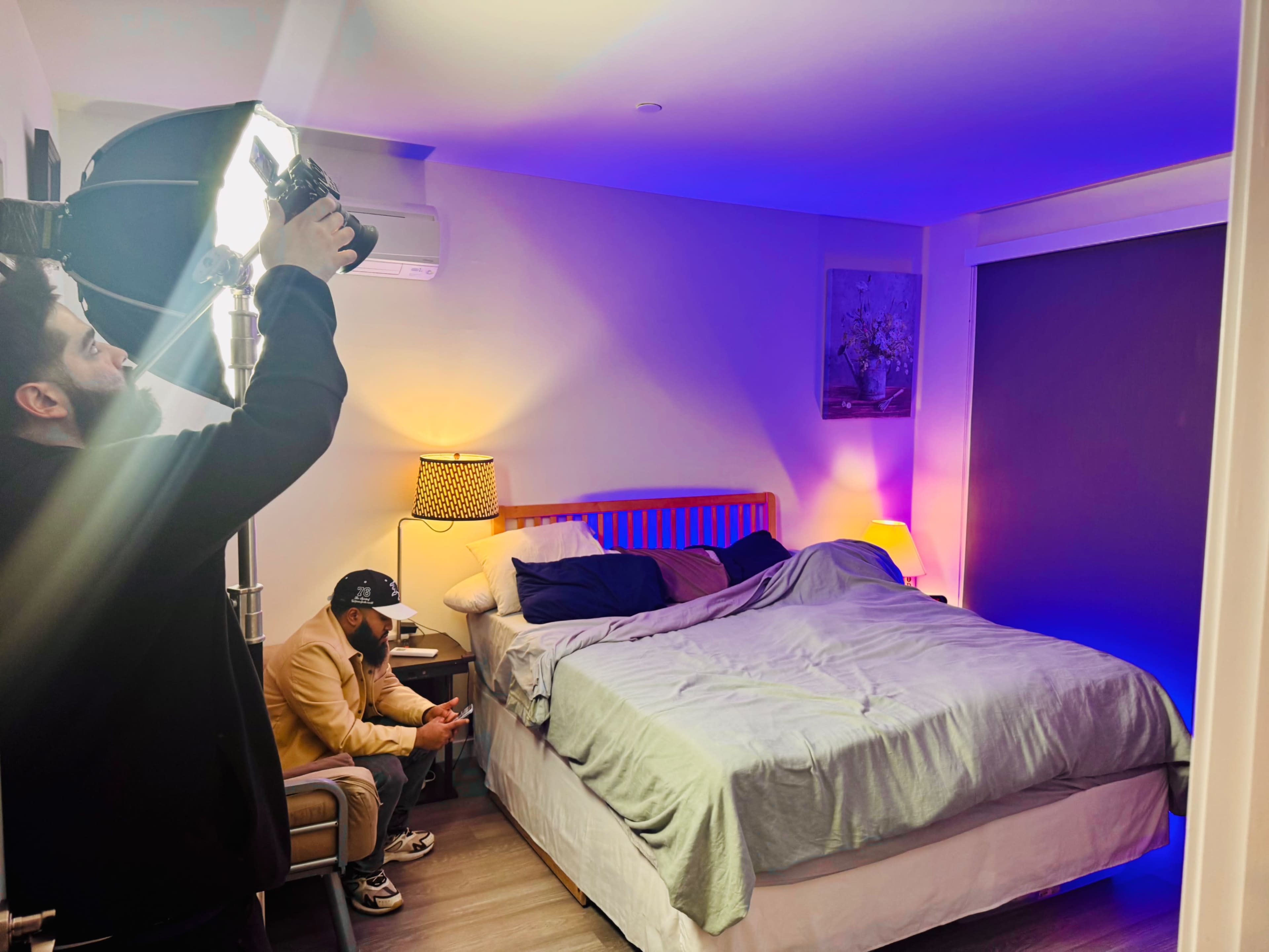 A person is adjusting a lighting setup above a bed in a softly lit bedroom while another person sits on a chair, looking at their phone.