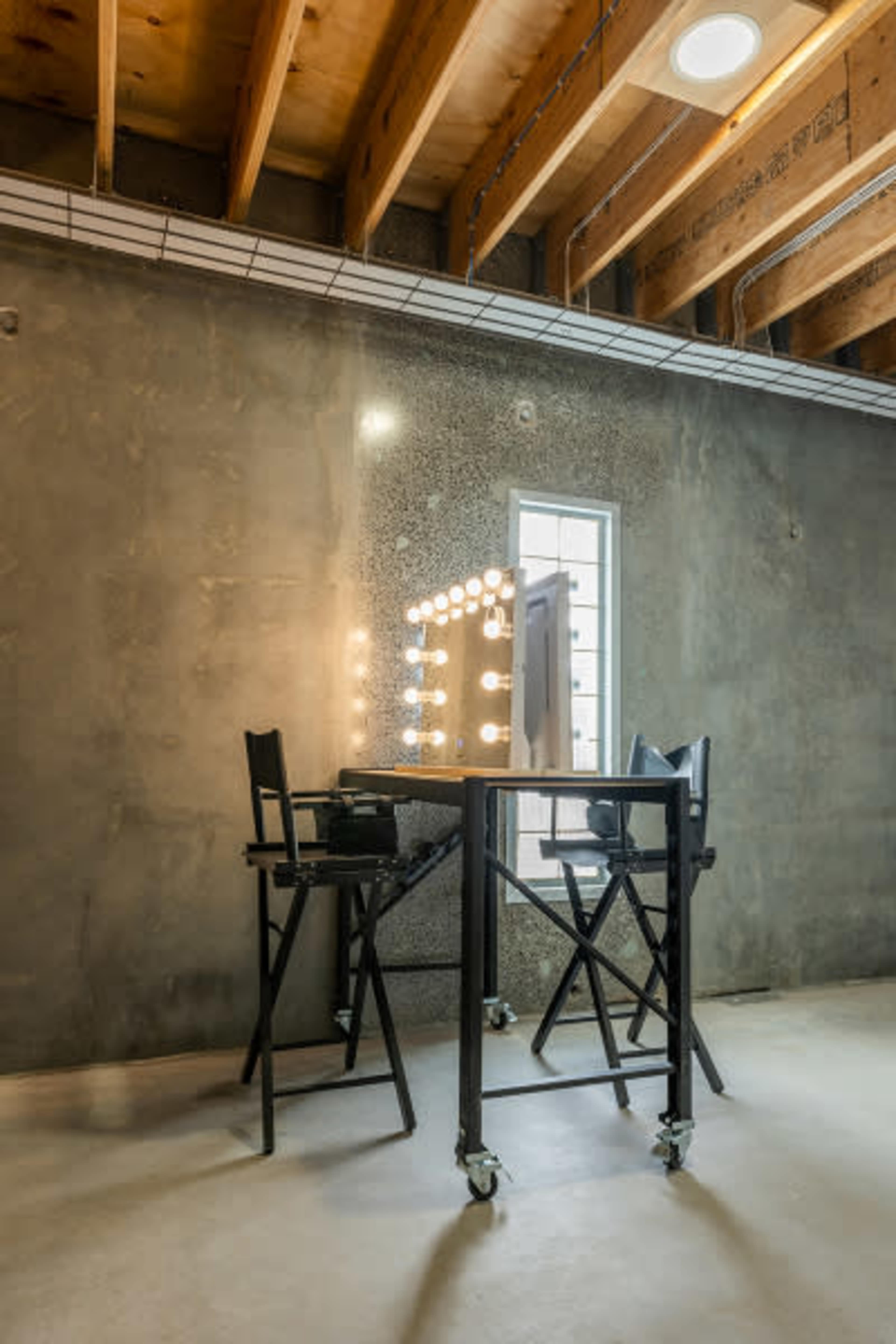 A makeup station with two black director's chairs and a large illuminated mirror against a concrete wall.