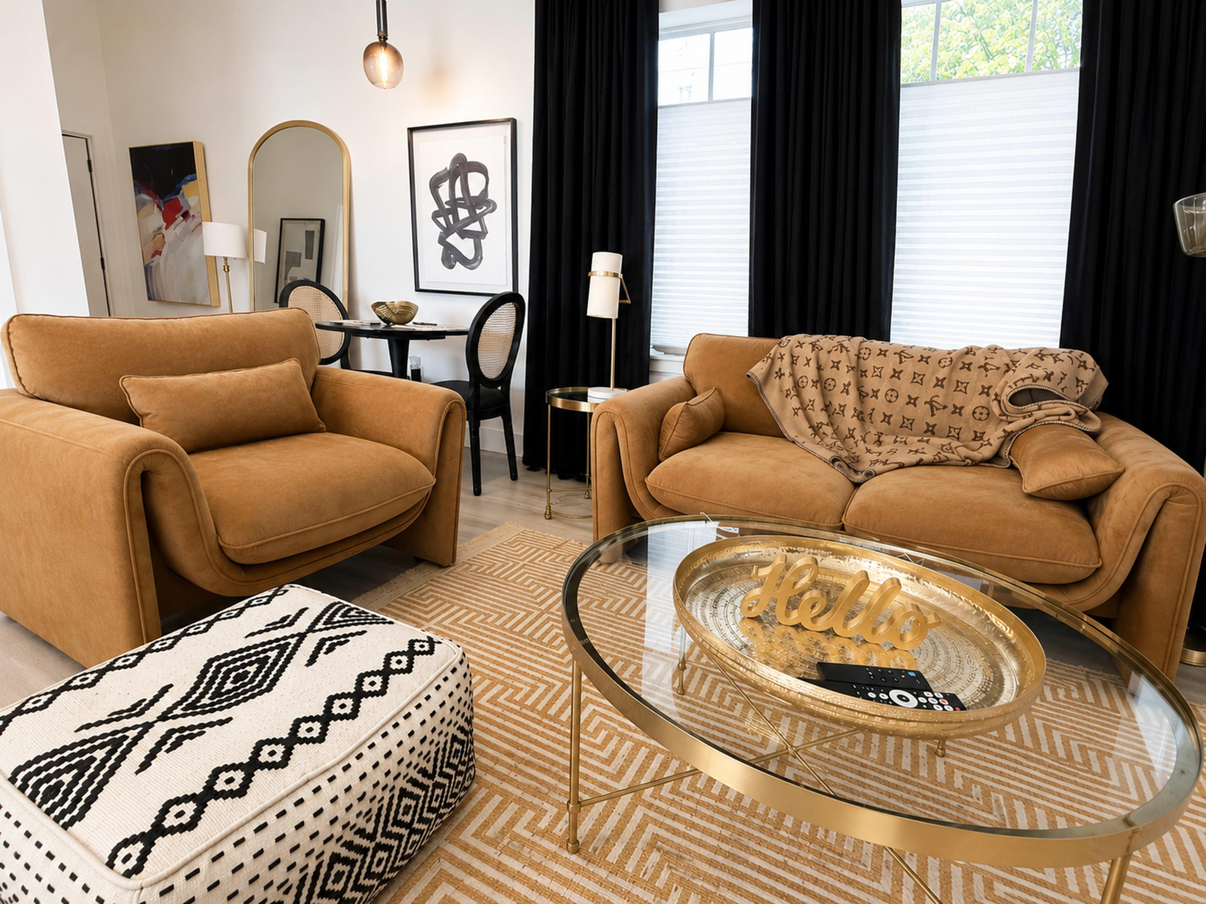 The image shows a cozy living room featuring two tan sofas, a glass coffee table with decorative items, and a patterned rug under a large window with sheer curtains.