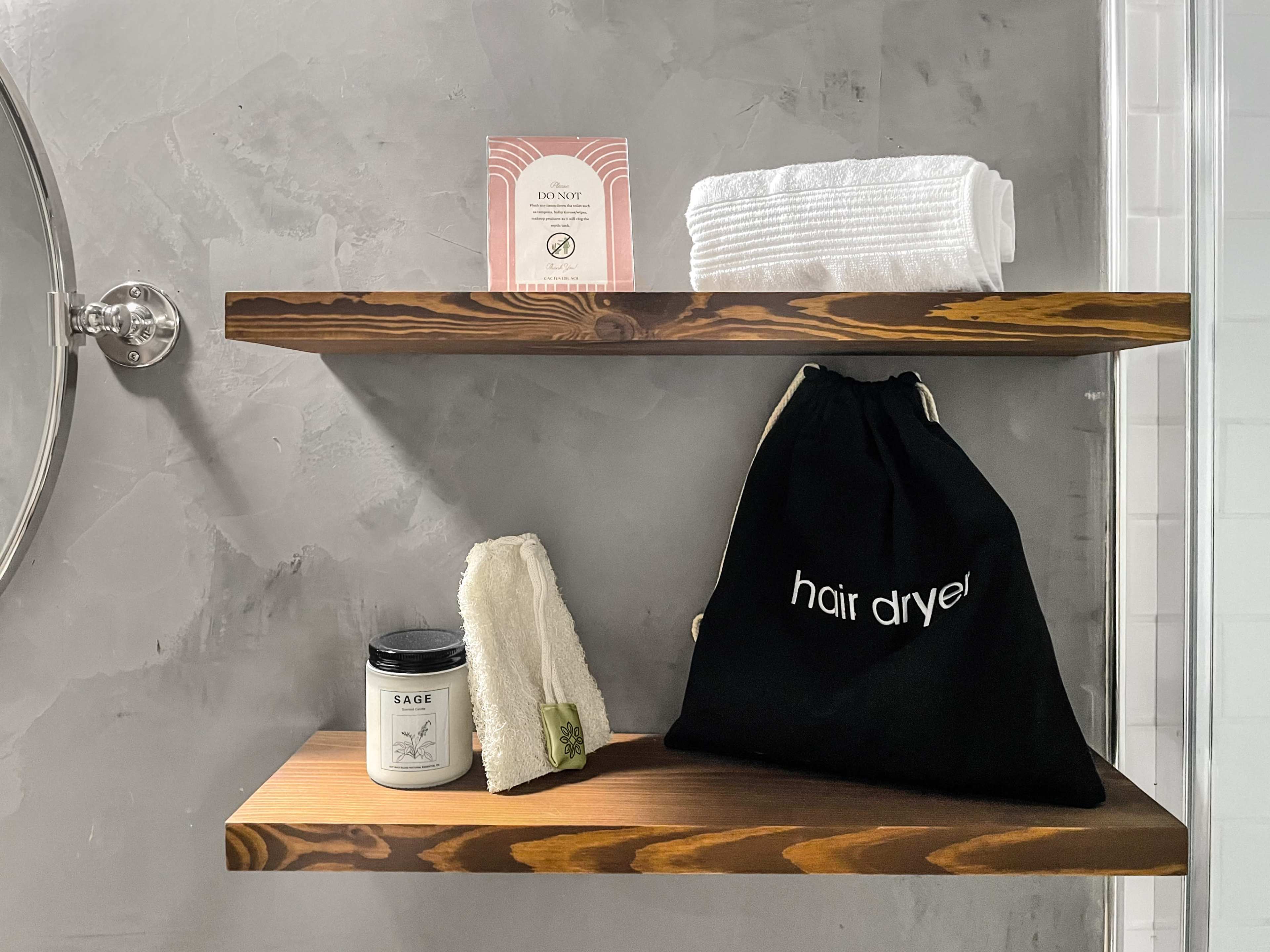 The image shows two wooden shelves in a bathroom with a pack of tissues, rolled towels, a scented candle, a washcloth, and a black bag labeled "hair dryer."