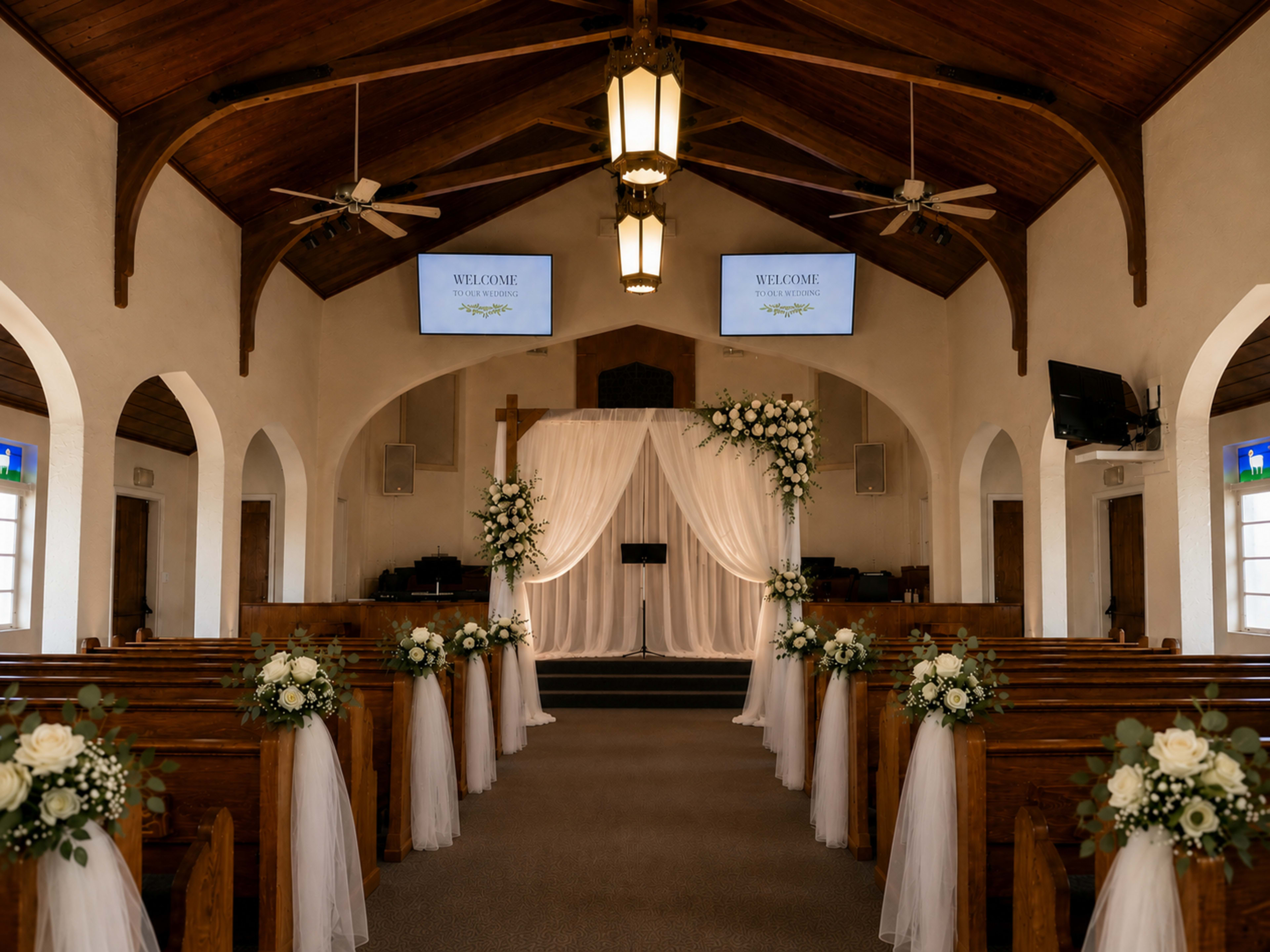 A wedding chapel features wooden beams, a central cross, and floral arrangements lining the pews, with screens displaying a welcome message at the front.