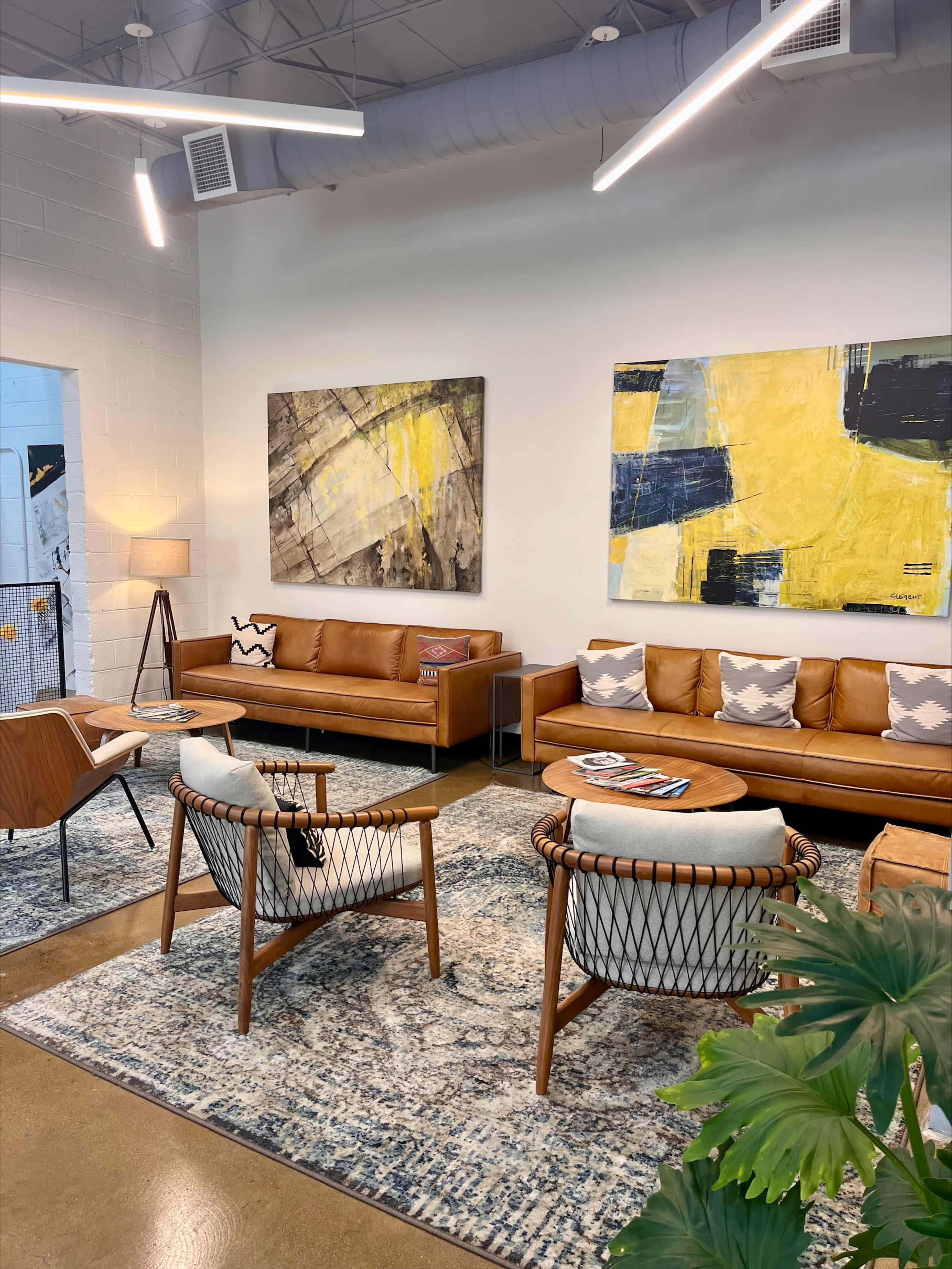A modern lounge area featuring brown leather sofas, two woven armchairs, a round coffee table with magazines, a patterned rug, and abstract artwork on the walls.