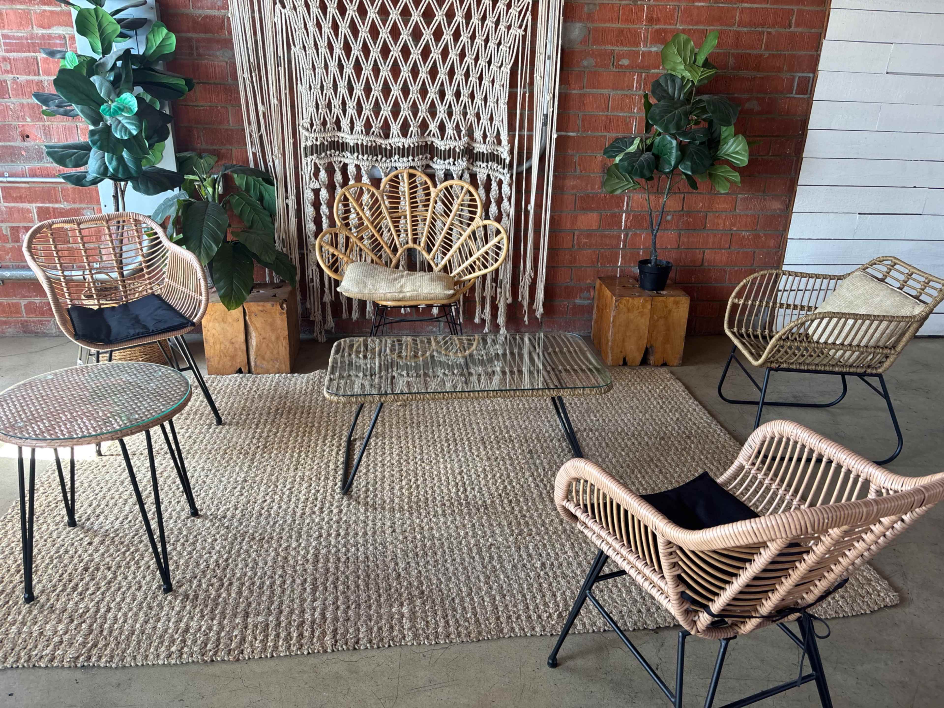 The image shows a cozy seating arrangement with five rattan chairs surrounding a glass coffee table, set on a woven rug against a brick wall with decorative plants.