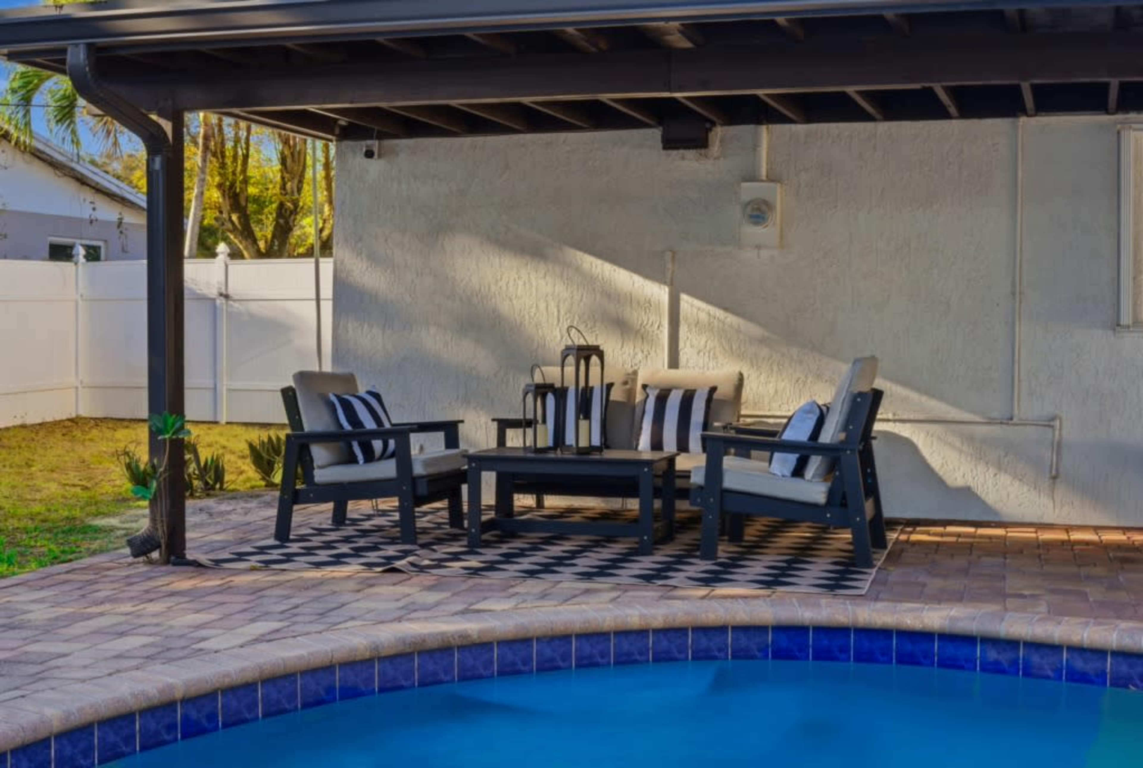 The image shows a patio area with comfortable seating, a coffee table, and a swimming pool adjacent to it.