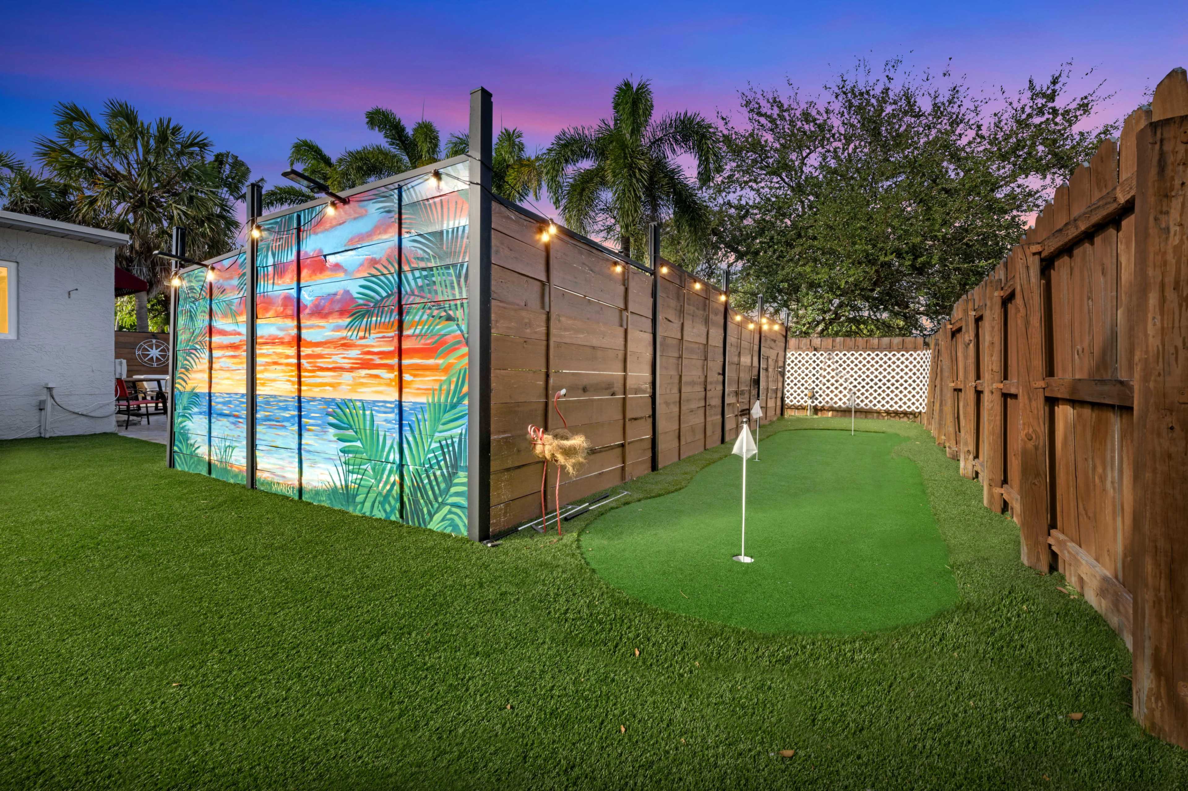 A small backyard with a putting green and a mural of a sunset over water on a wooden fence.