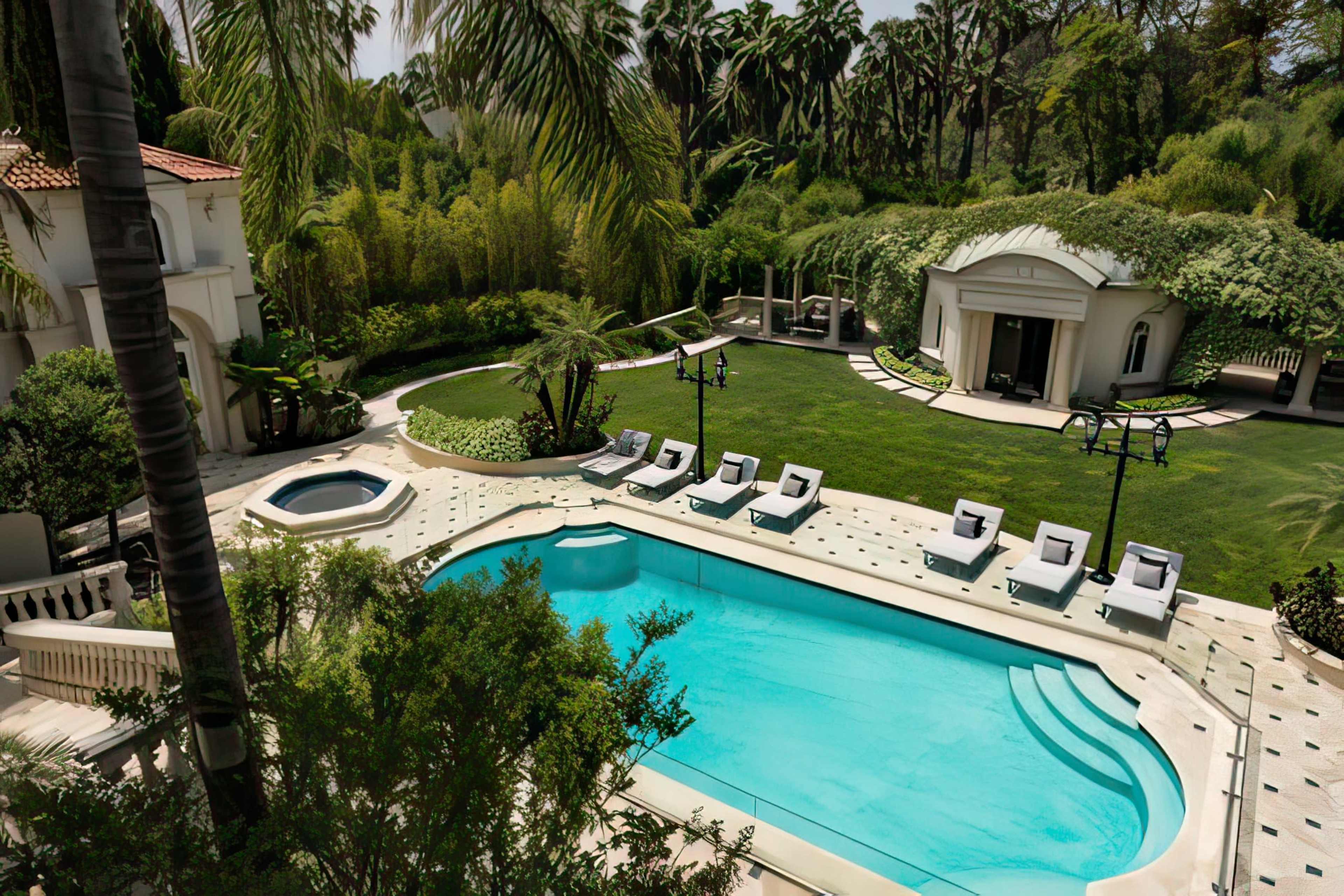 The image showcases a luxurious backyard featuring a large swimming pool, a spa, lounge chairs, and a manicured garden surrounded by palm trees.