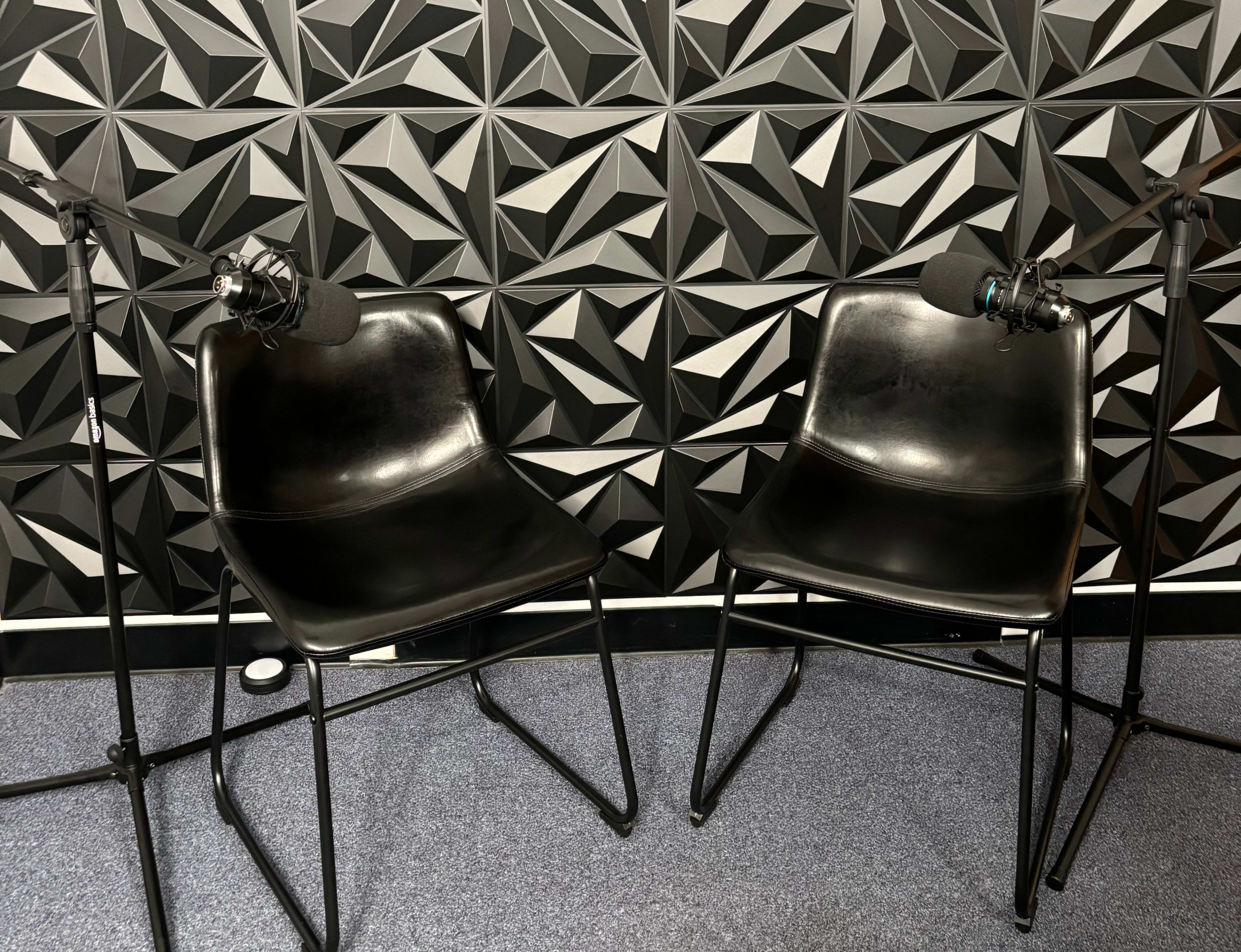 Two black chairs are positioned facing each other, each equipped with a microphone on a stand, against a textured black wall.