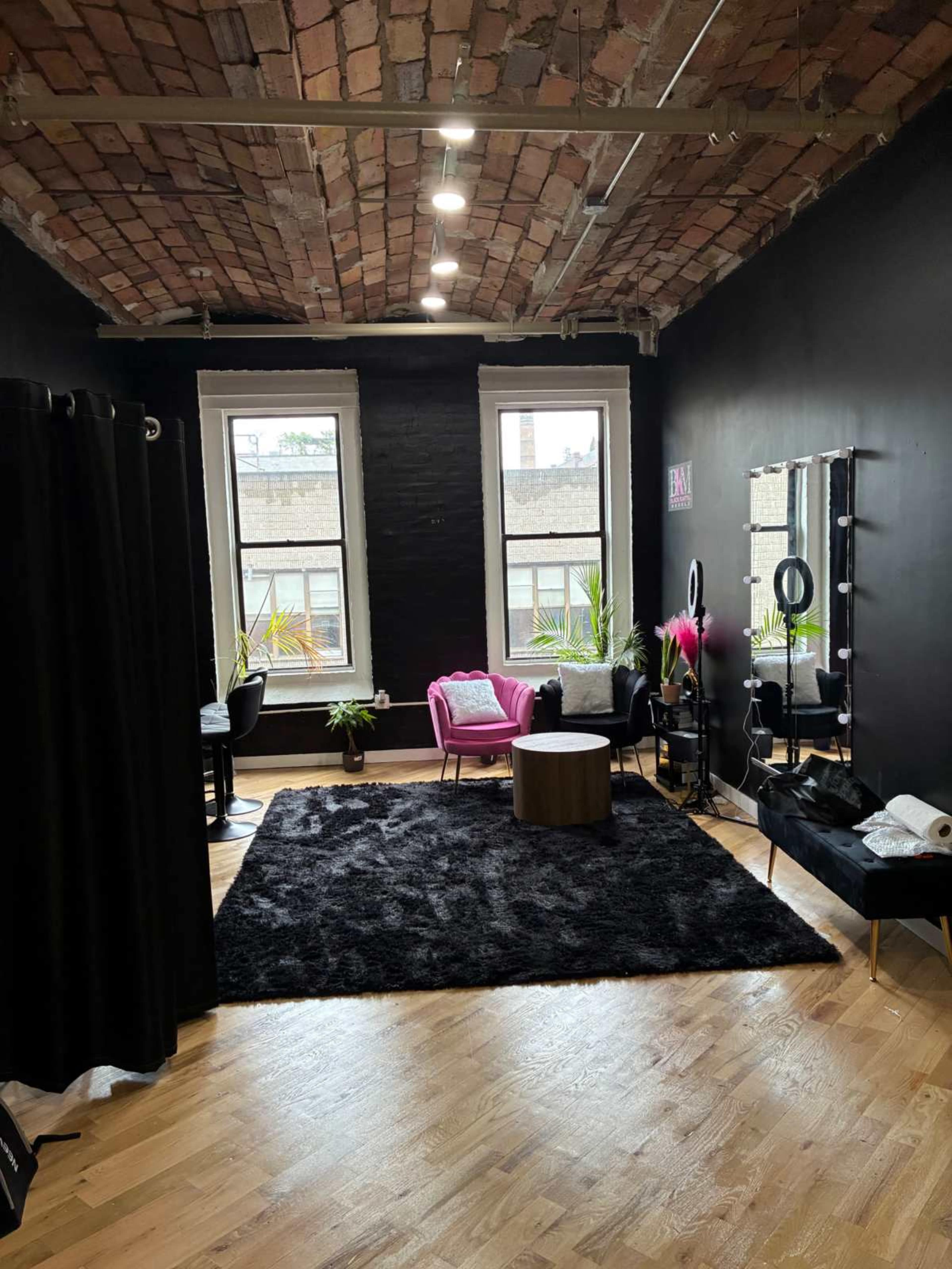 The image shows a modern studio space with black walls, a brick ceiling, large windows, and a plush black rug, featuring a pink chair, a wooden table, and a vanity with lights.