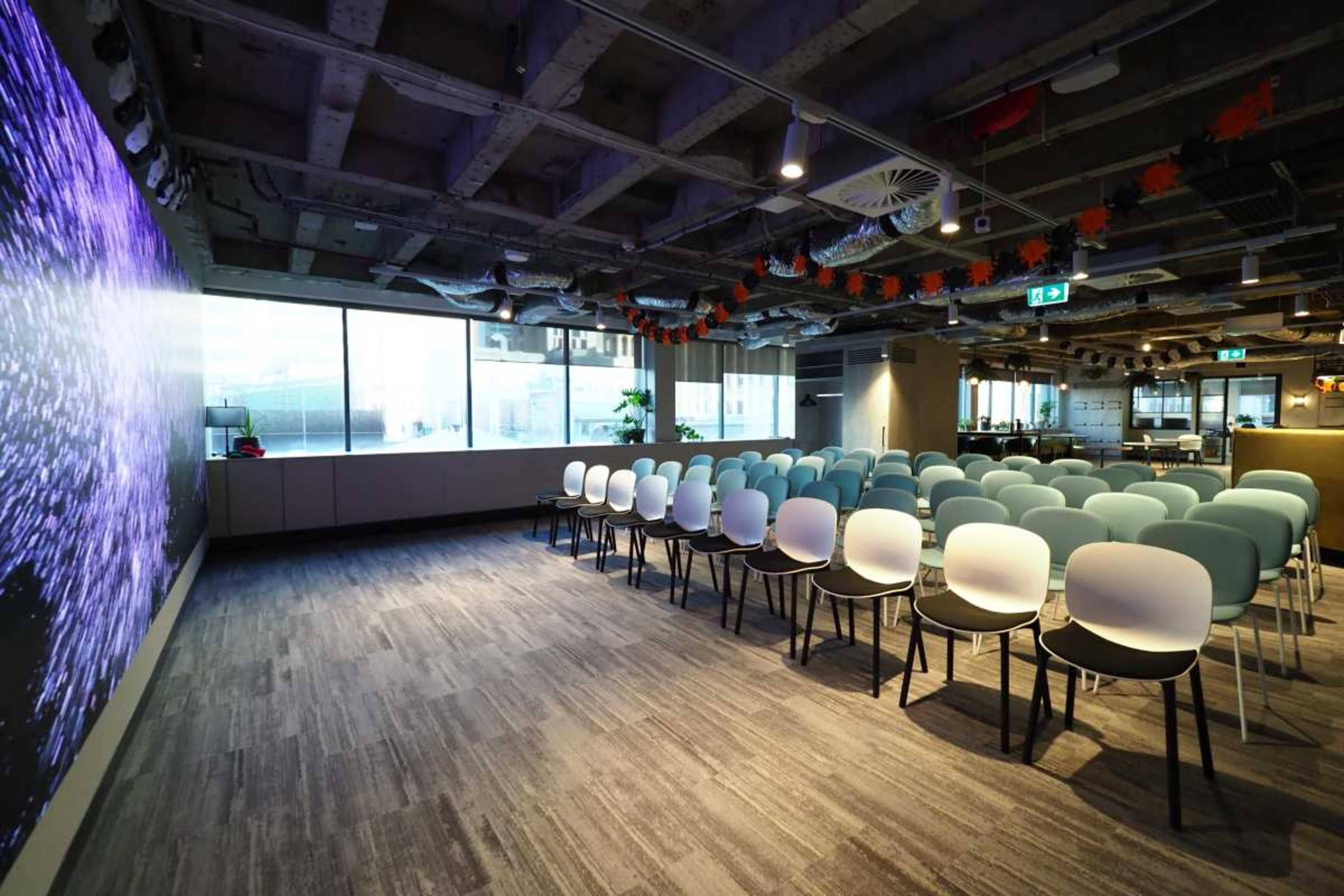 The image shows a modern conference room set up with rows of chairs facing a large screen, under industrial-style ceiling elements and decorative accents.