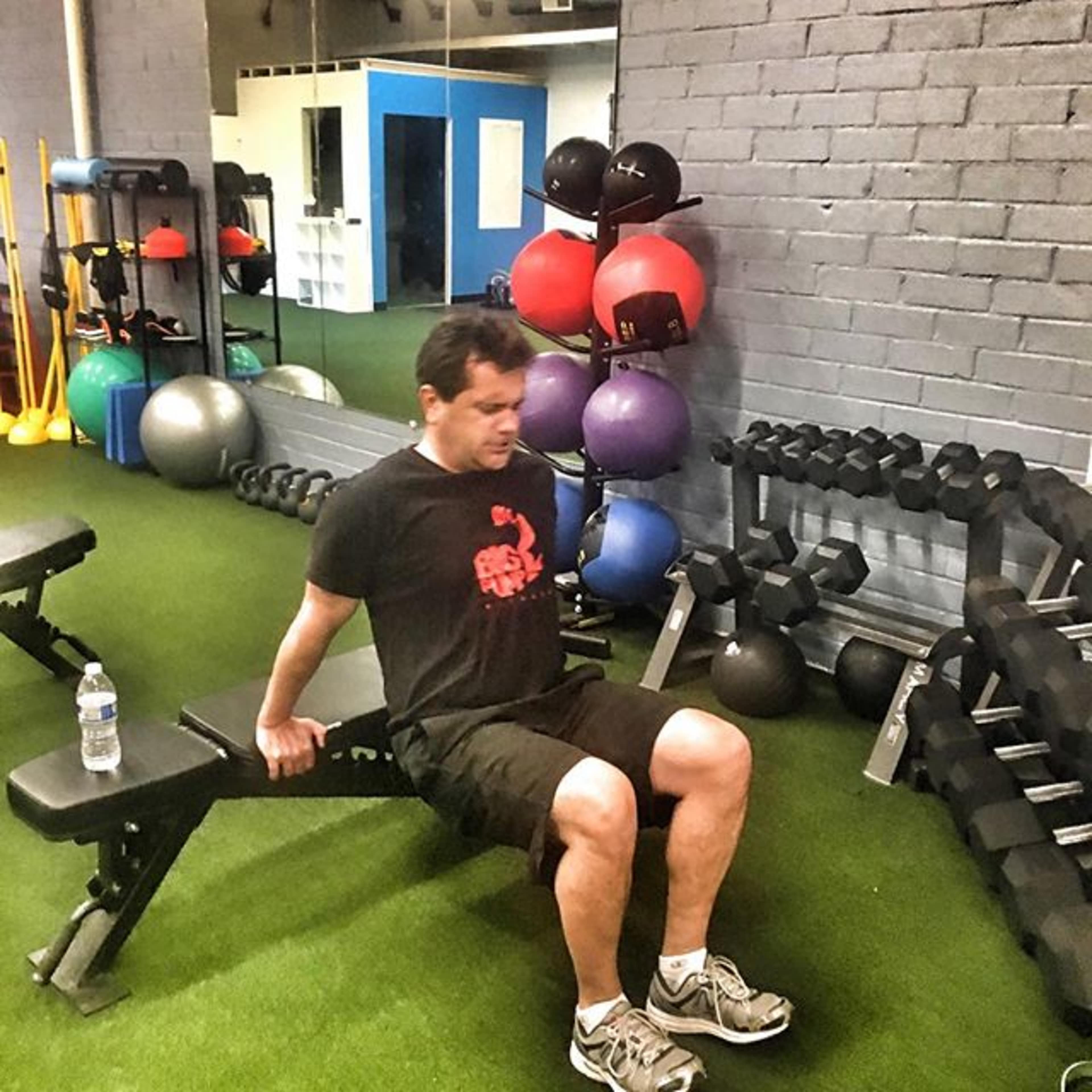 A man performs a tricep dip on a bench in a gym with various exercise equipment around him.