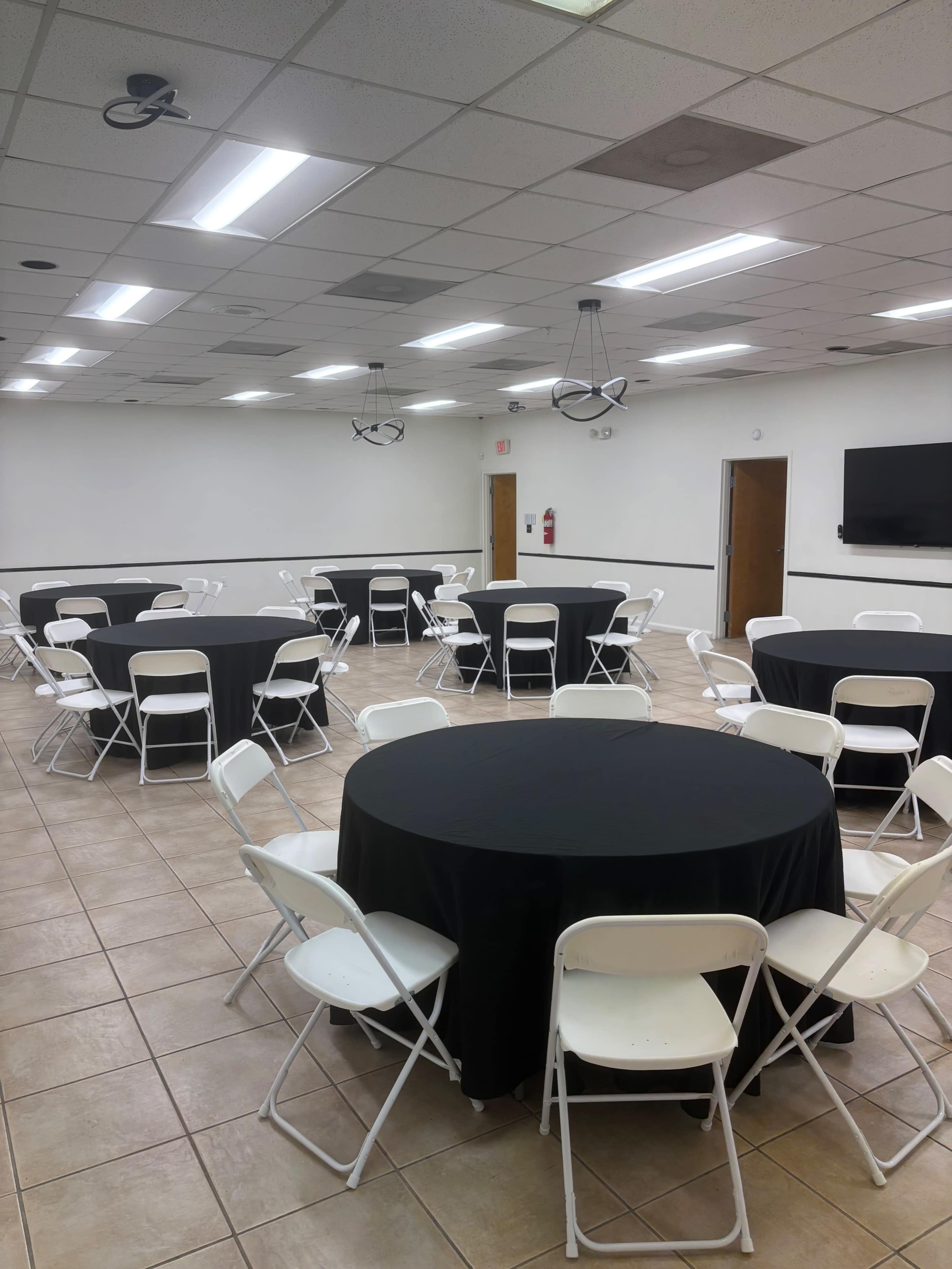 The image shows a large room with multiple round tables covered in black tablecloths, each surrounded by white chairs, and bright overhead lighting.