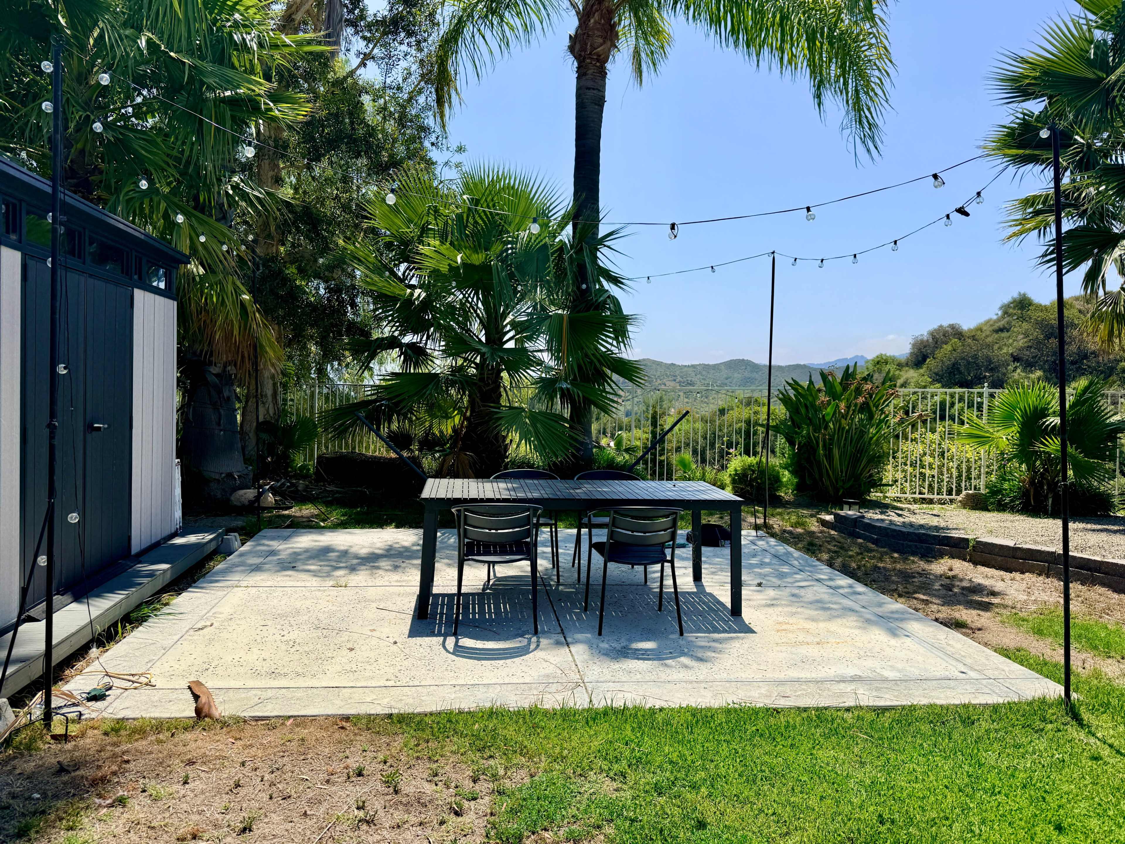 A dining table with four chairs is set on a concrete patio surrounded by palm trees and greenery under a clear blue sky.