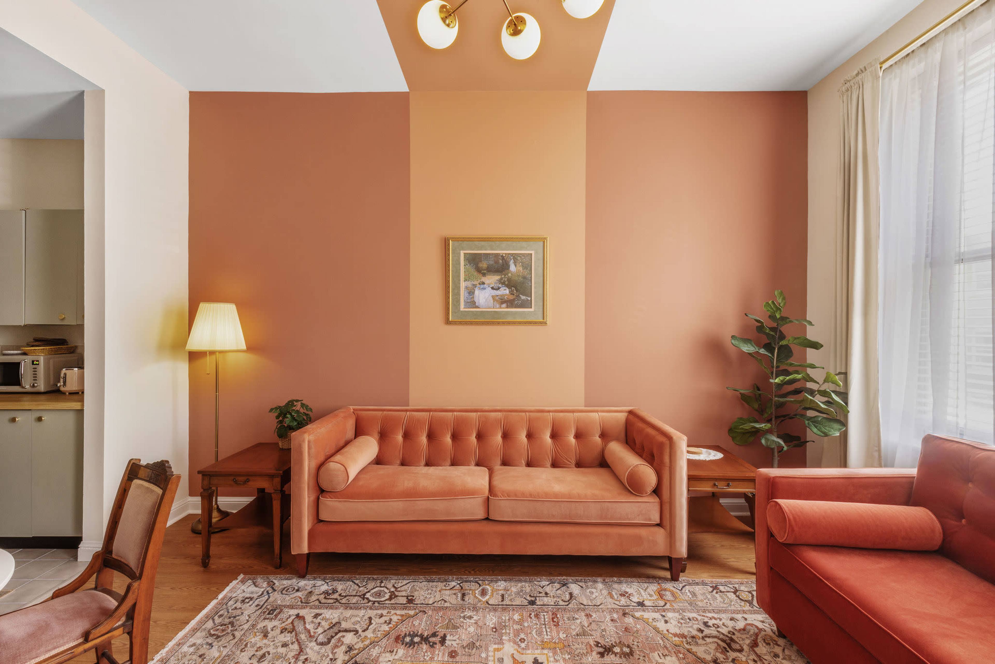 The image shows a living room with a sofa and armchair in warm tones, a painting on the wall, a floor lamp, and a potted plant.