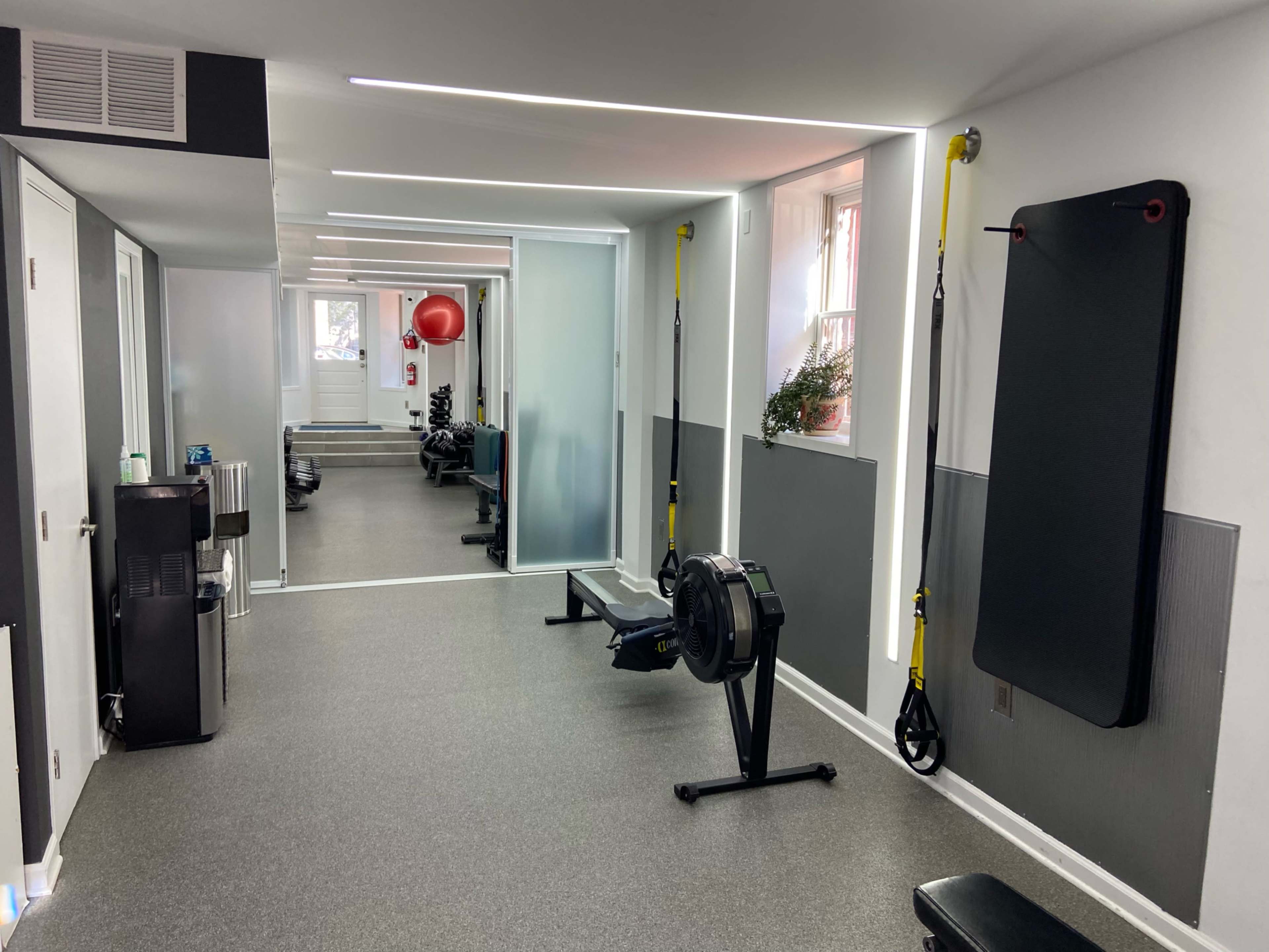 A modern gym interior with exercise equipment including a rowing machine and wall-mounted TRX straps, illuminated by overhead lights.