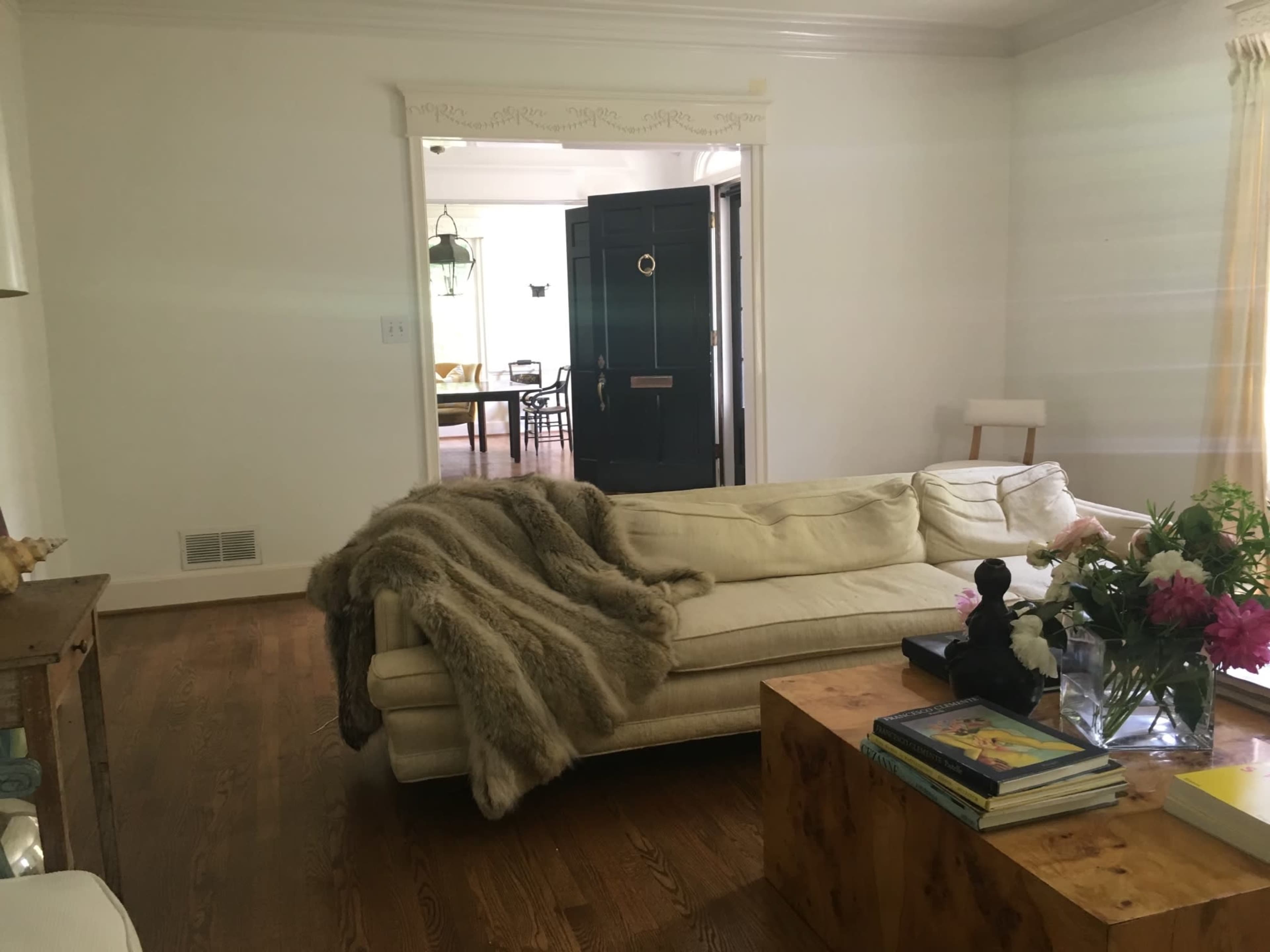 A cozy living room with a white sofa draped with a fur throw, a wooden coffee table, and a vase of flowers, alongside an entrance door leading to another room.