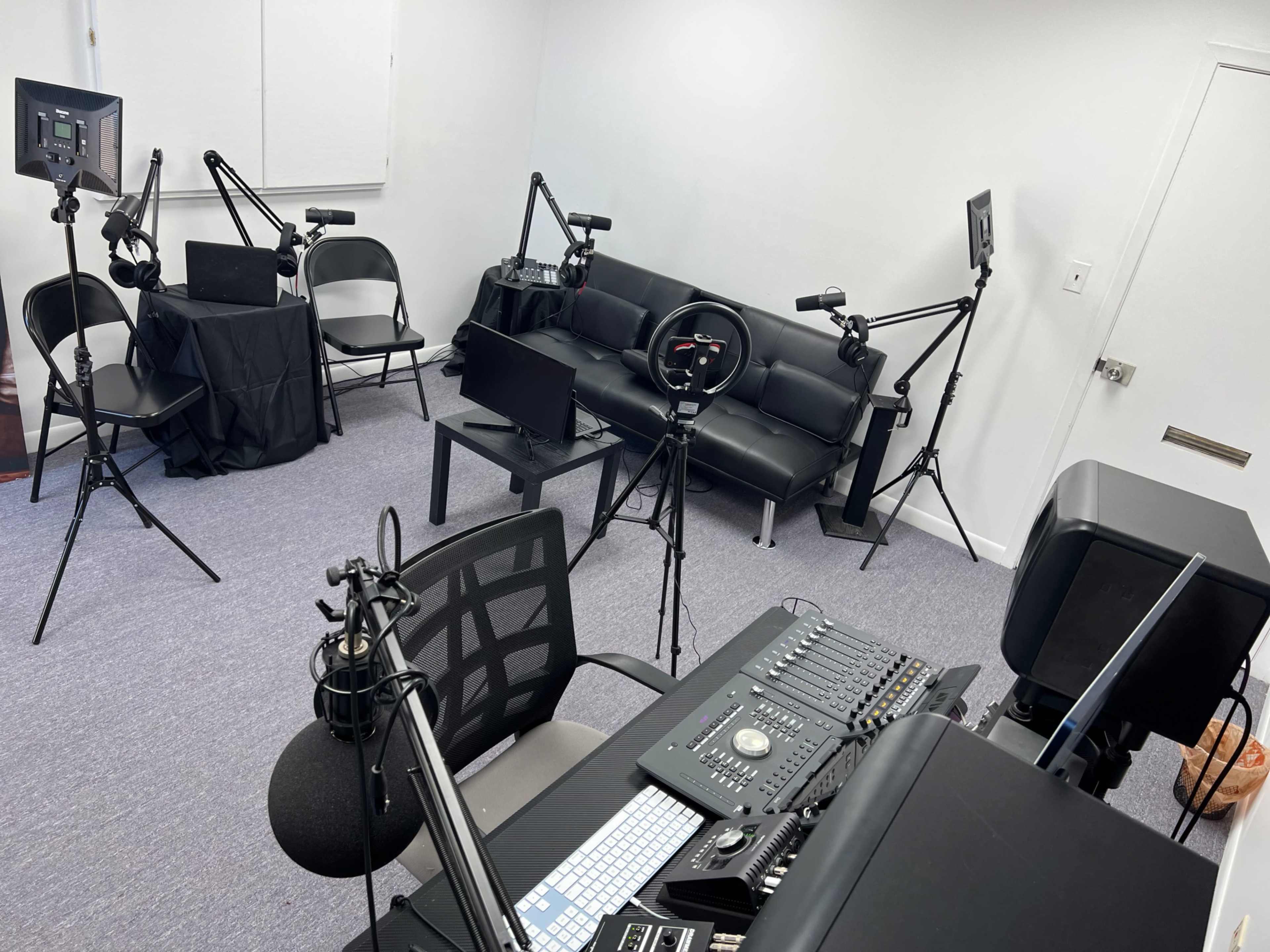 A small studio setup with multiple recording equipment, a black couch, and chairs arranged around a central table.