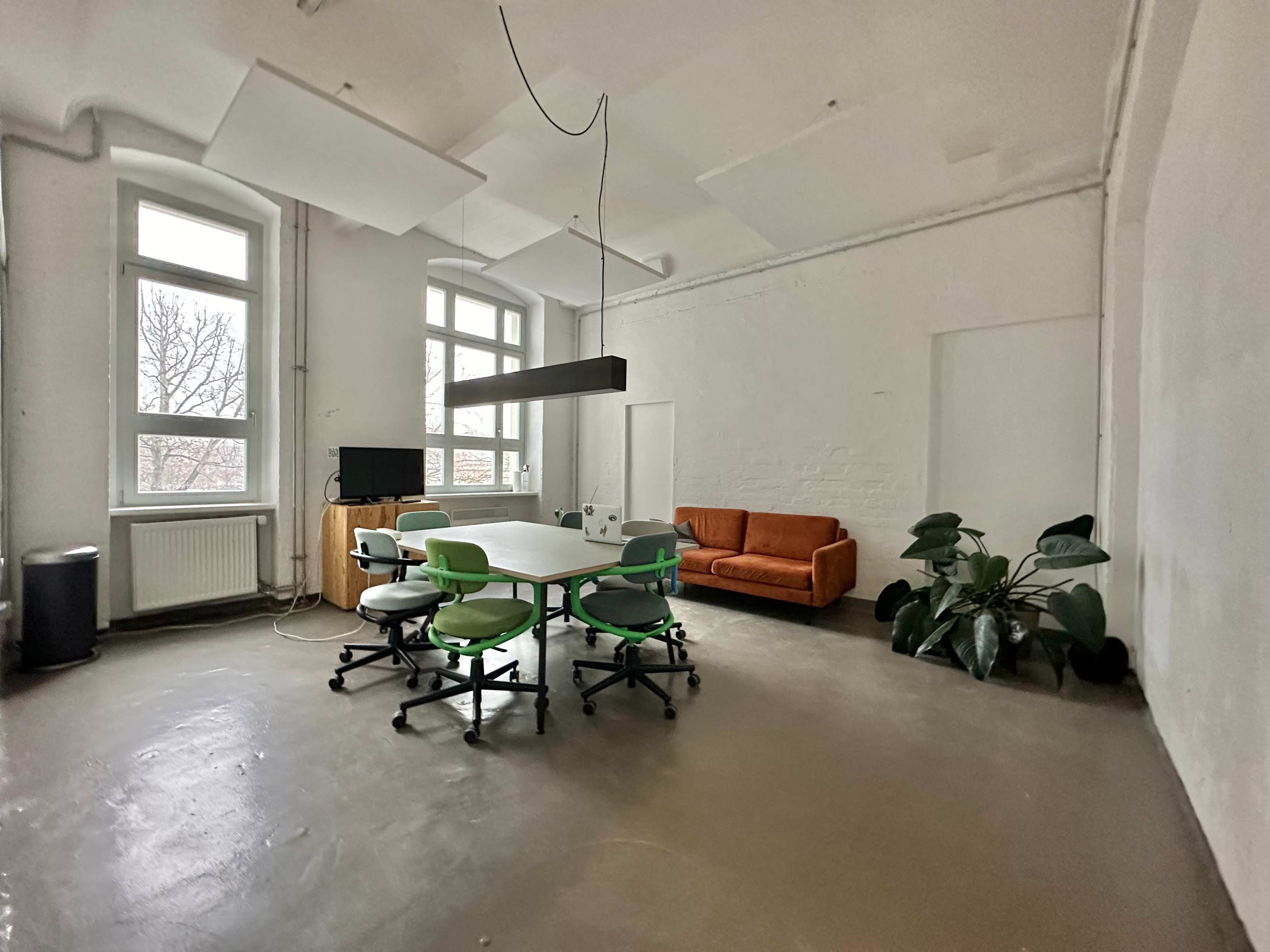 A spacious room features a central meeting table surrounded by green office chairs, an orange sofa, a television, and large windows allowing natural light.