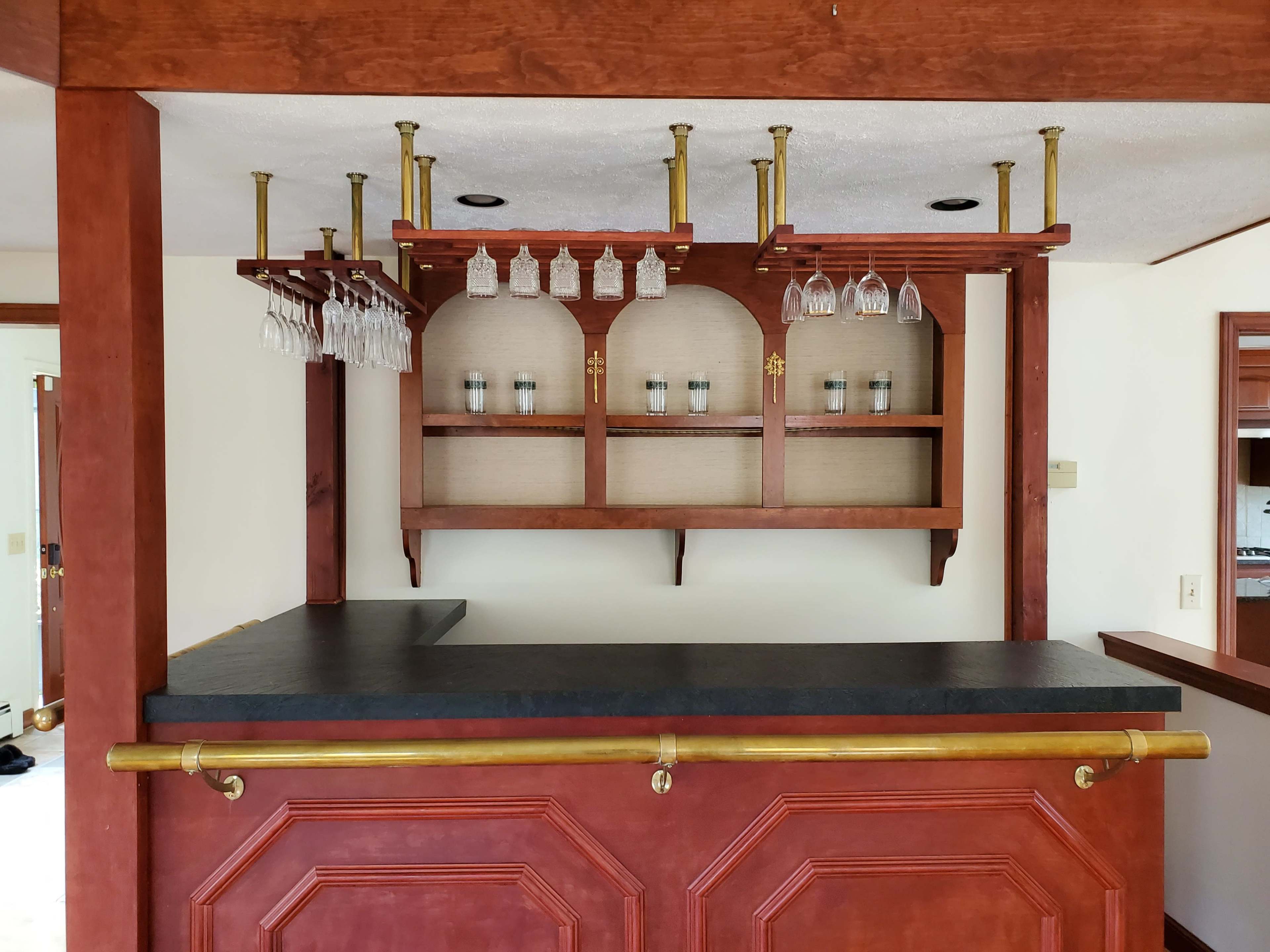 A wooden bar with a dark countertop and hanging glassware, complemented by a shelf displaying jars in an interior space.
