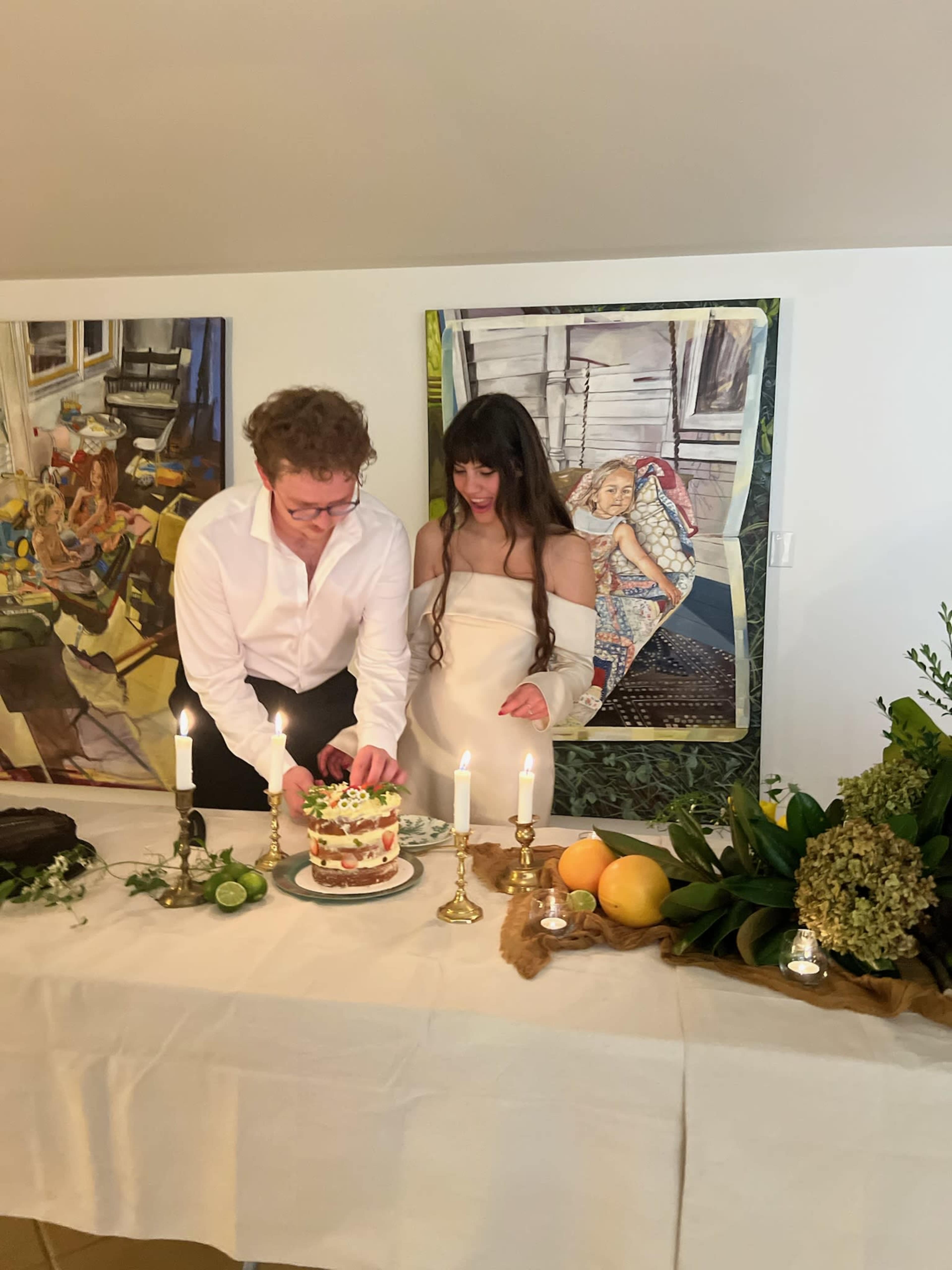 A man and a woman are cutting a cake together in front of a decorated table with two candles and various green plants.