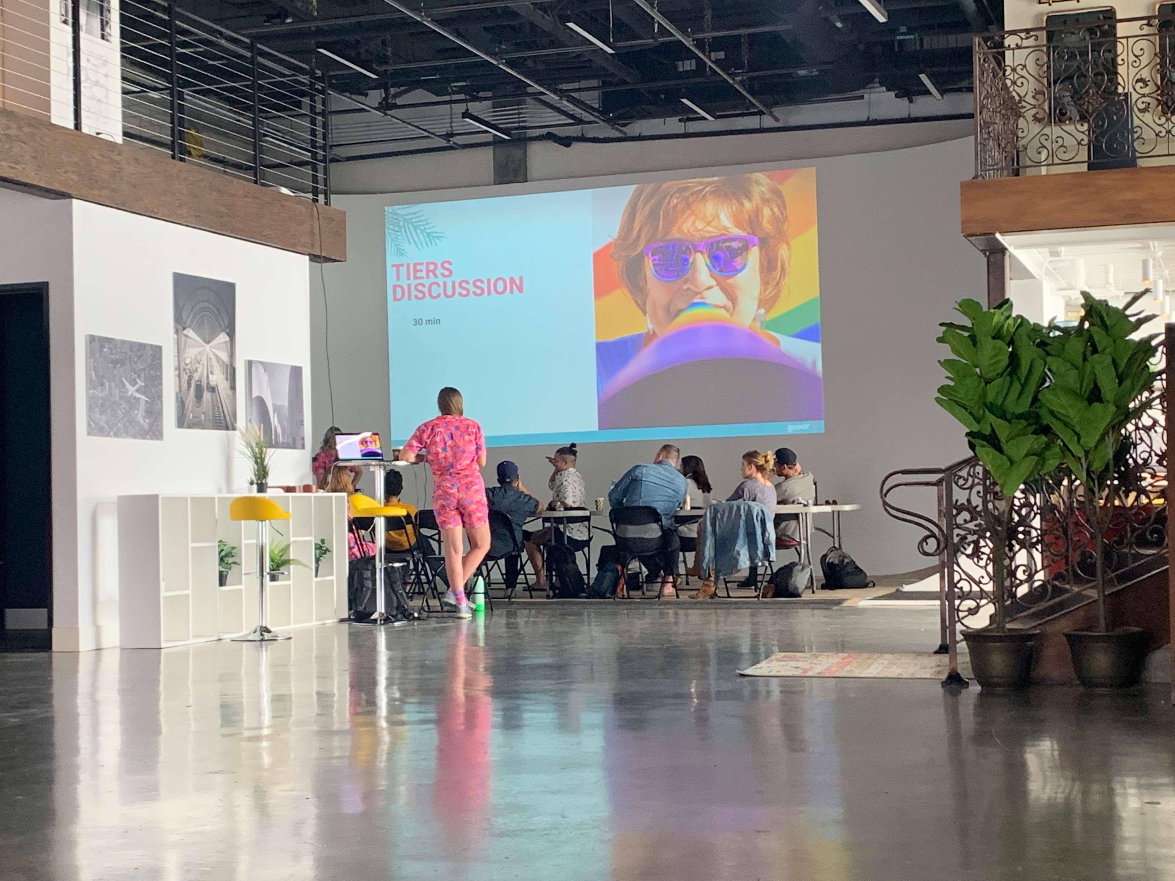 A group of people sits at a table in a well-lit room, with a large screen displaying the words "TIERS DISCUSSION" alongside a colorful image.
