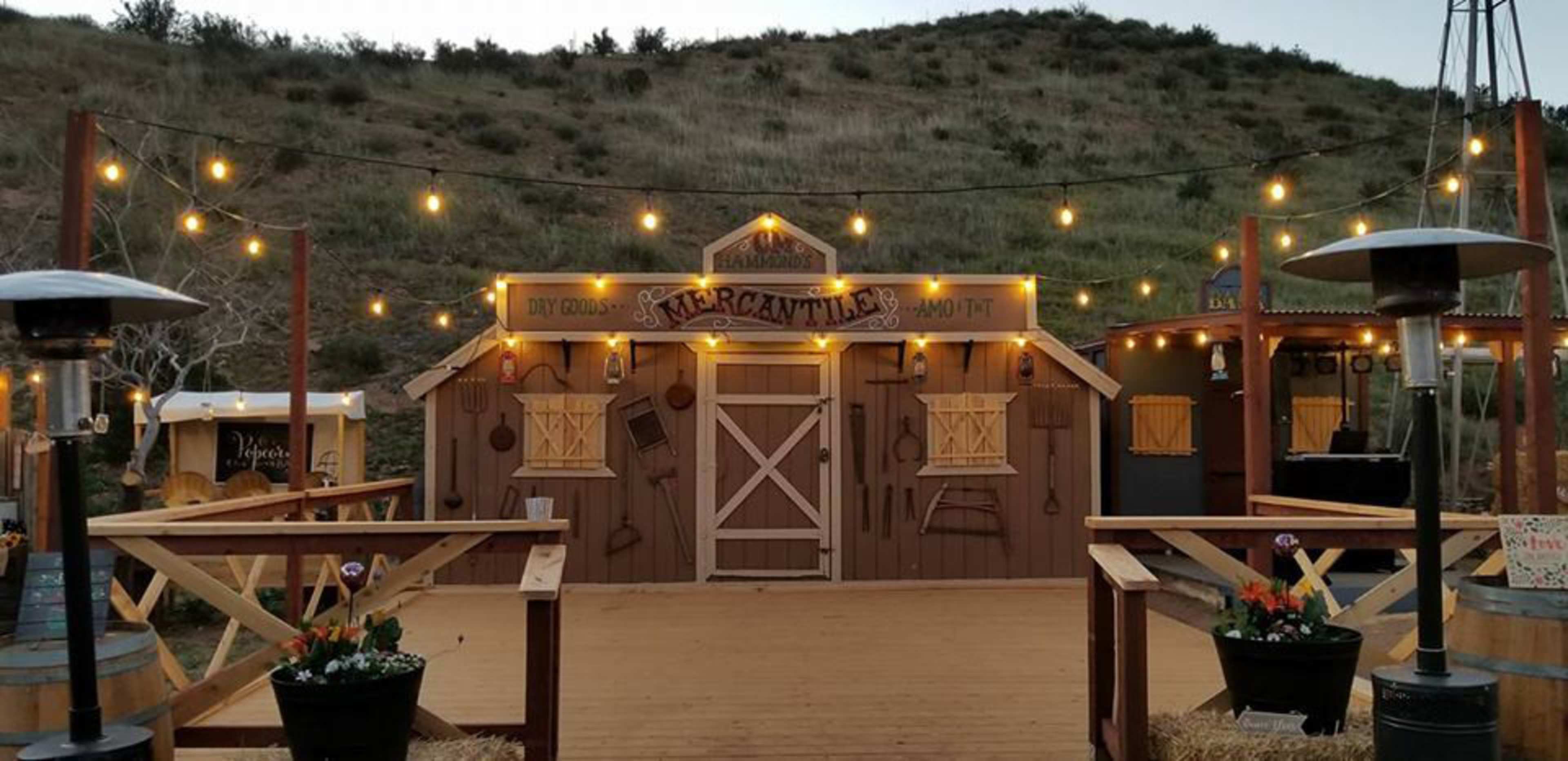 A rustic building with a wooden facade, illuminated by string lights, situated in a rural outdoor setting with a hillside in the background.