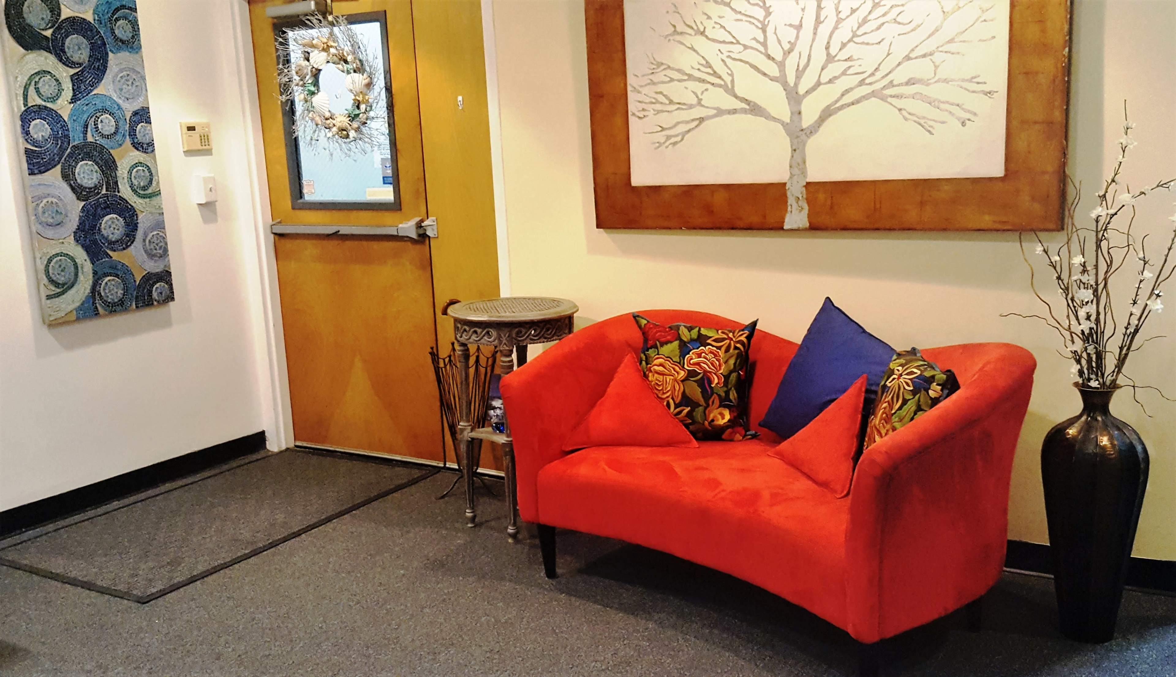 A red curved sofa with colorful patterned cushions is positioned next to a decorative table and a tall vase in a room featuring artwork on the walls.