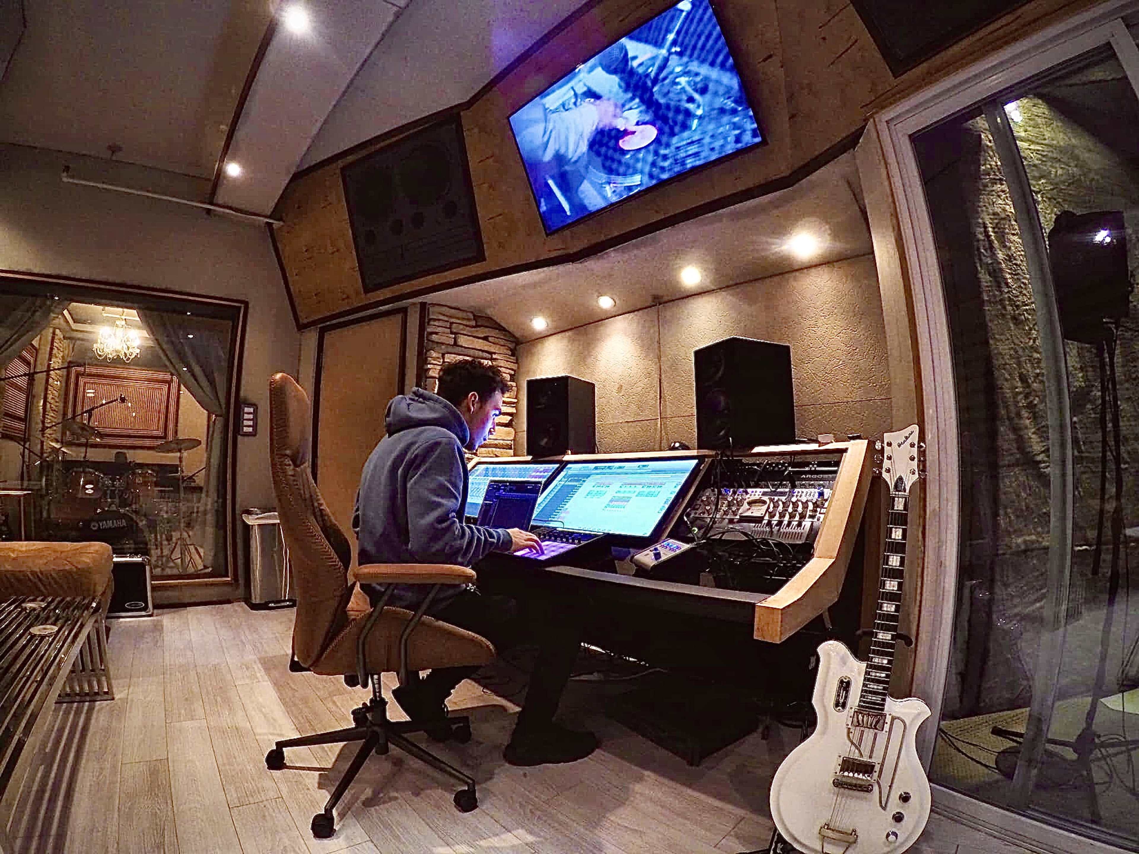 A person sits at a large audio mixing console in a sound studio, working on a computer with monitors displaying music software.