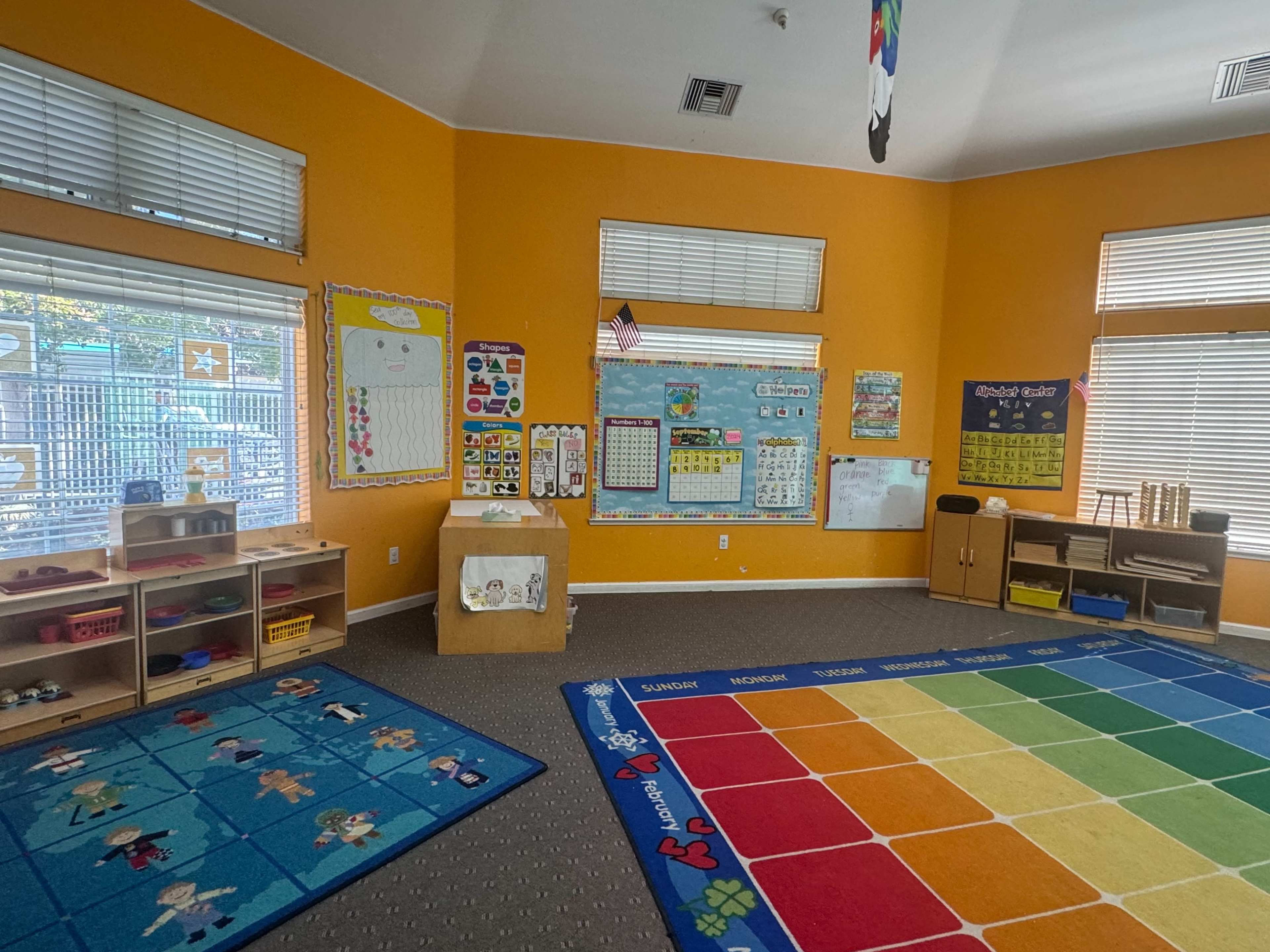 The image shows a brightly colored classroom with orange walls, a carpet with divided colored squares, and educational posters on the walls.