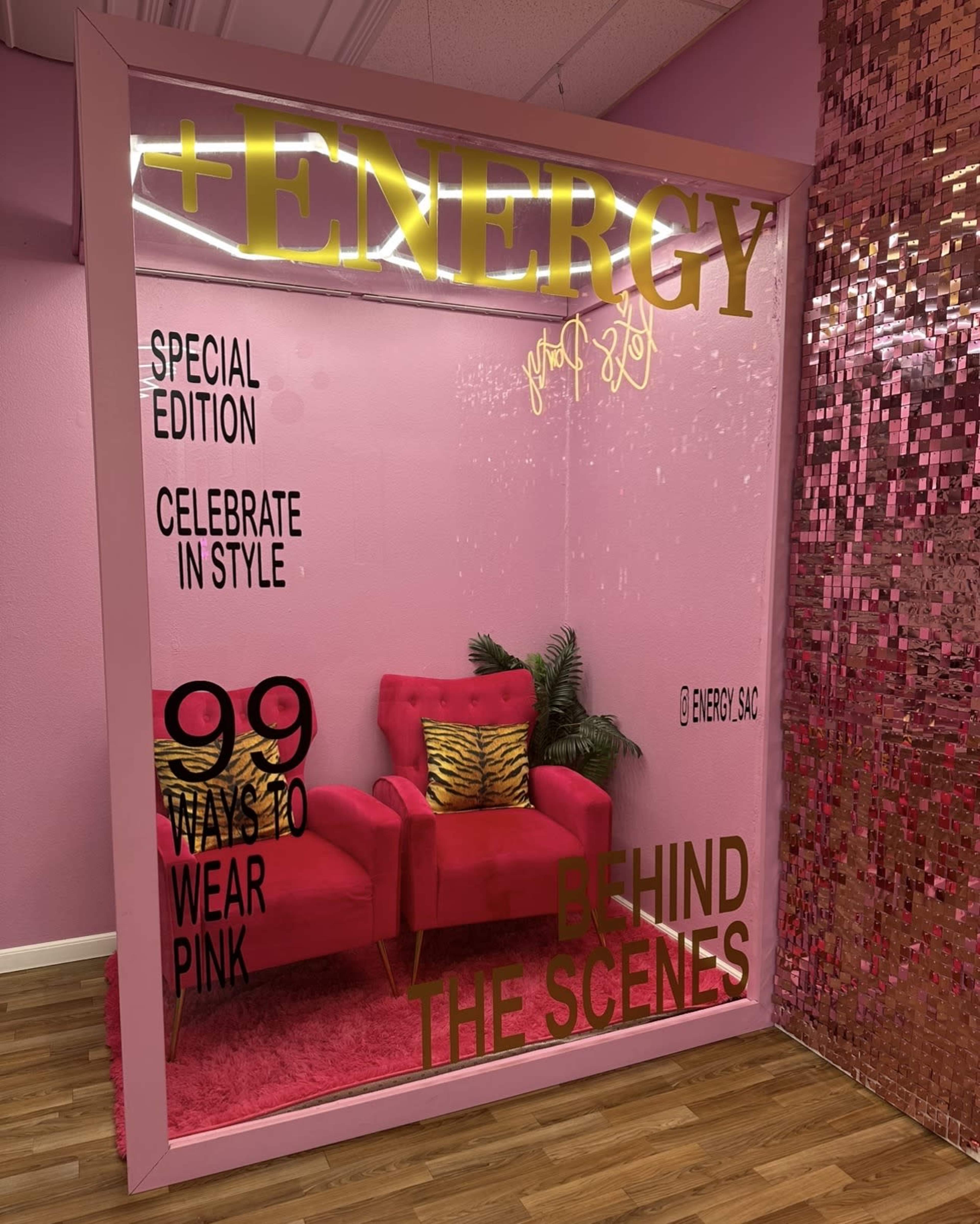 The image depicts a vibrant pink-themed display featuring a cozy seating area with a pink couch, adorned with a decorative backdrop and promotional text highlighting a special event.