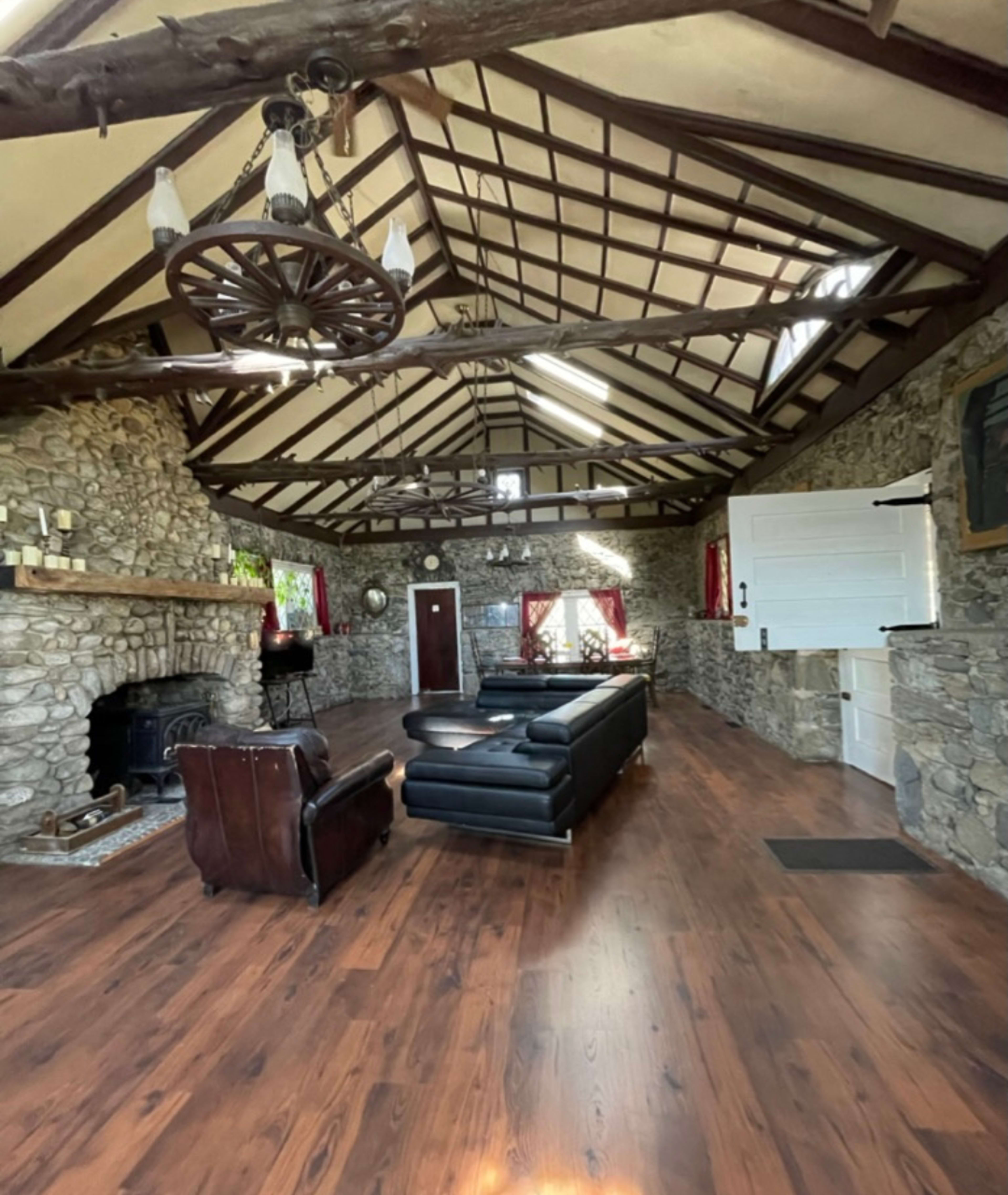 The image showcases a spacious living room featuring stone walls, a high wooden ceiling with beams, and a black leather sofa centered on a wooden floor.