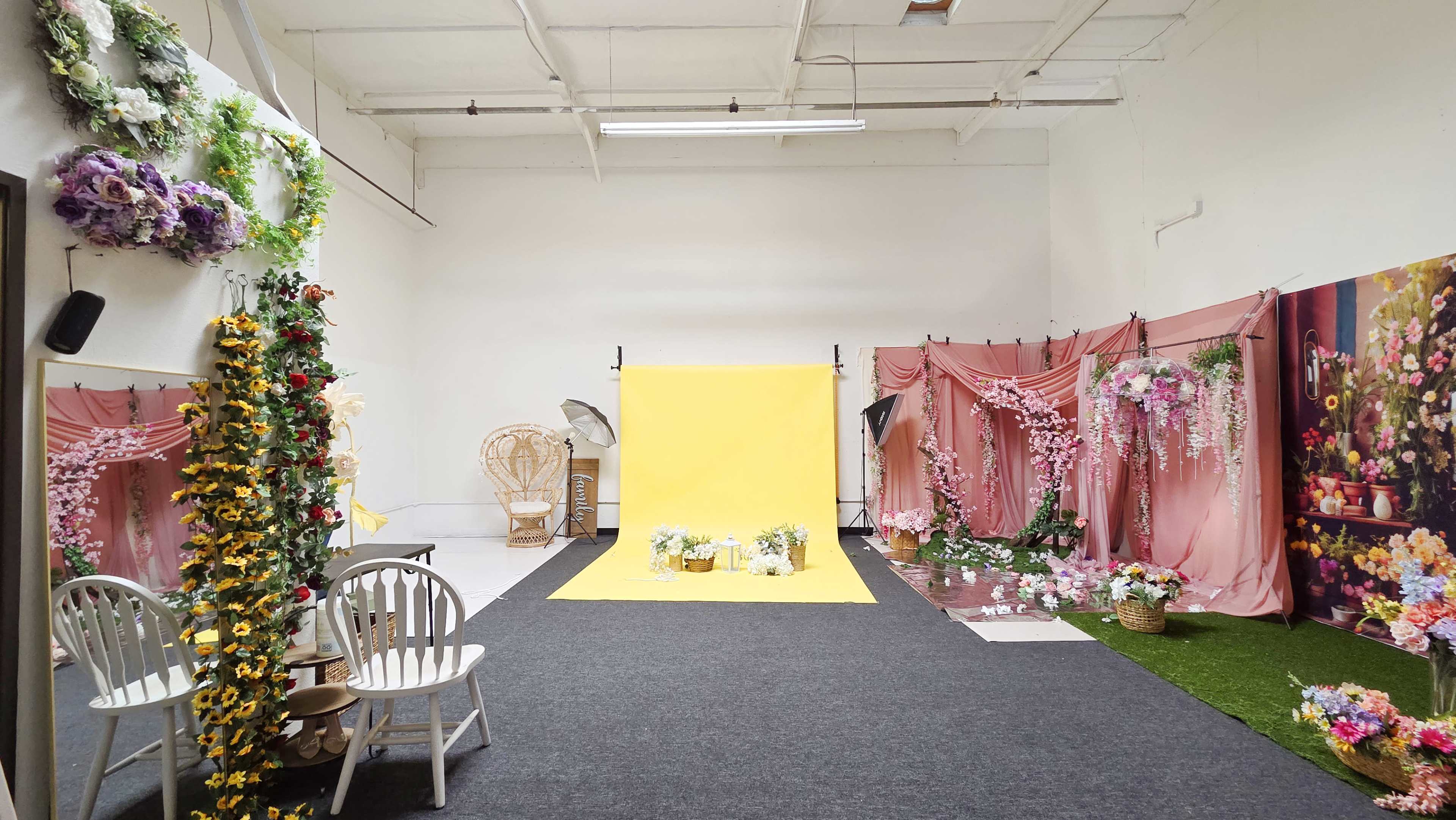 The image shows a spacious photo studio featuring a yellow backdrop, a floral arrangement, pink drapery, and decorative elements including chairs and artificial flowers.
