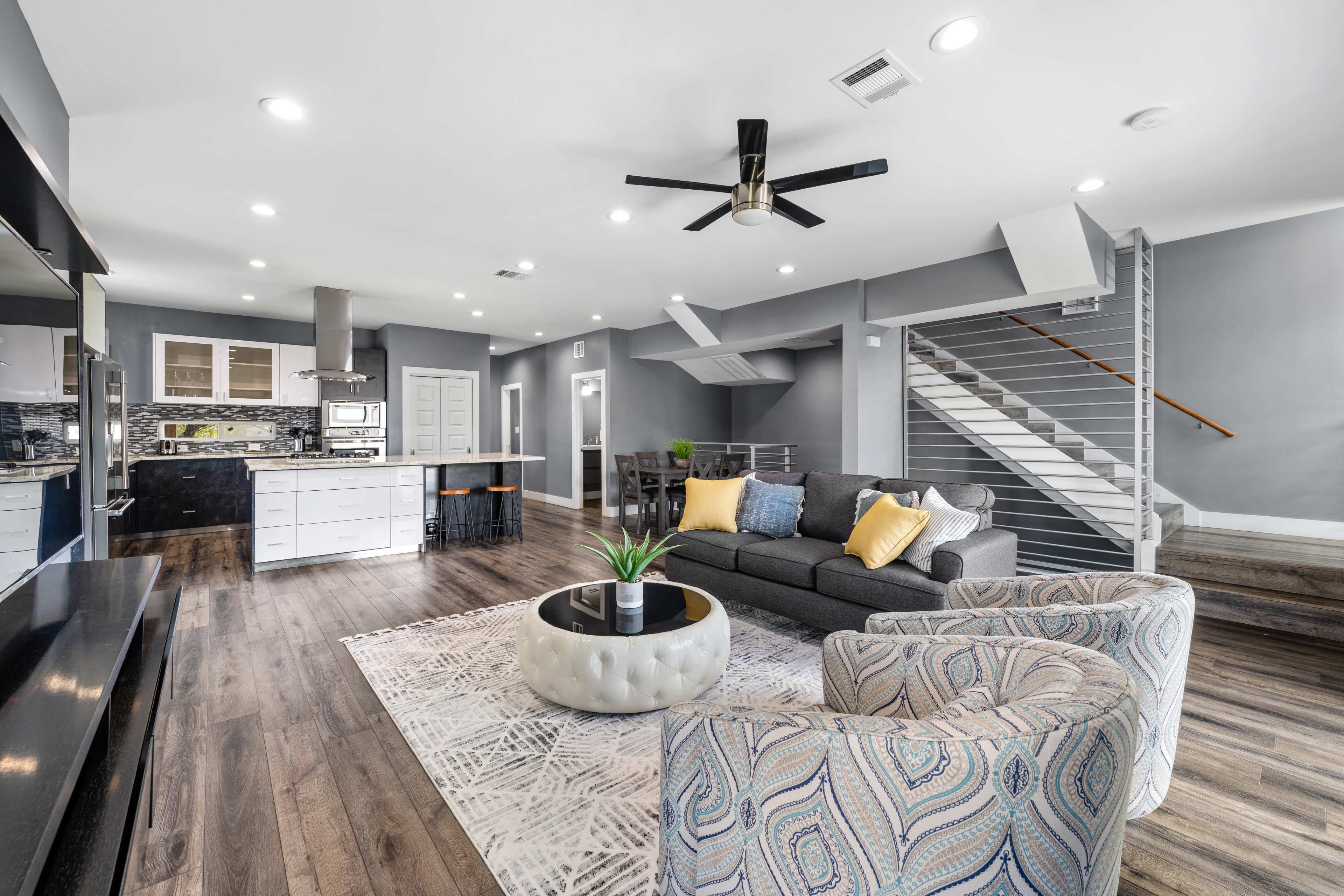 A modern living space featuring a gray sectional sofa, two patterned chairs, and a coffee table, with a staircase and kitchen area in the background.