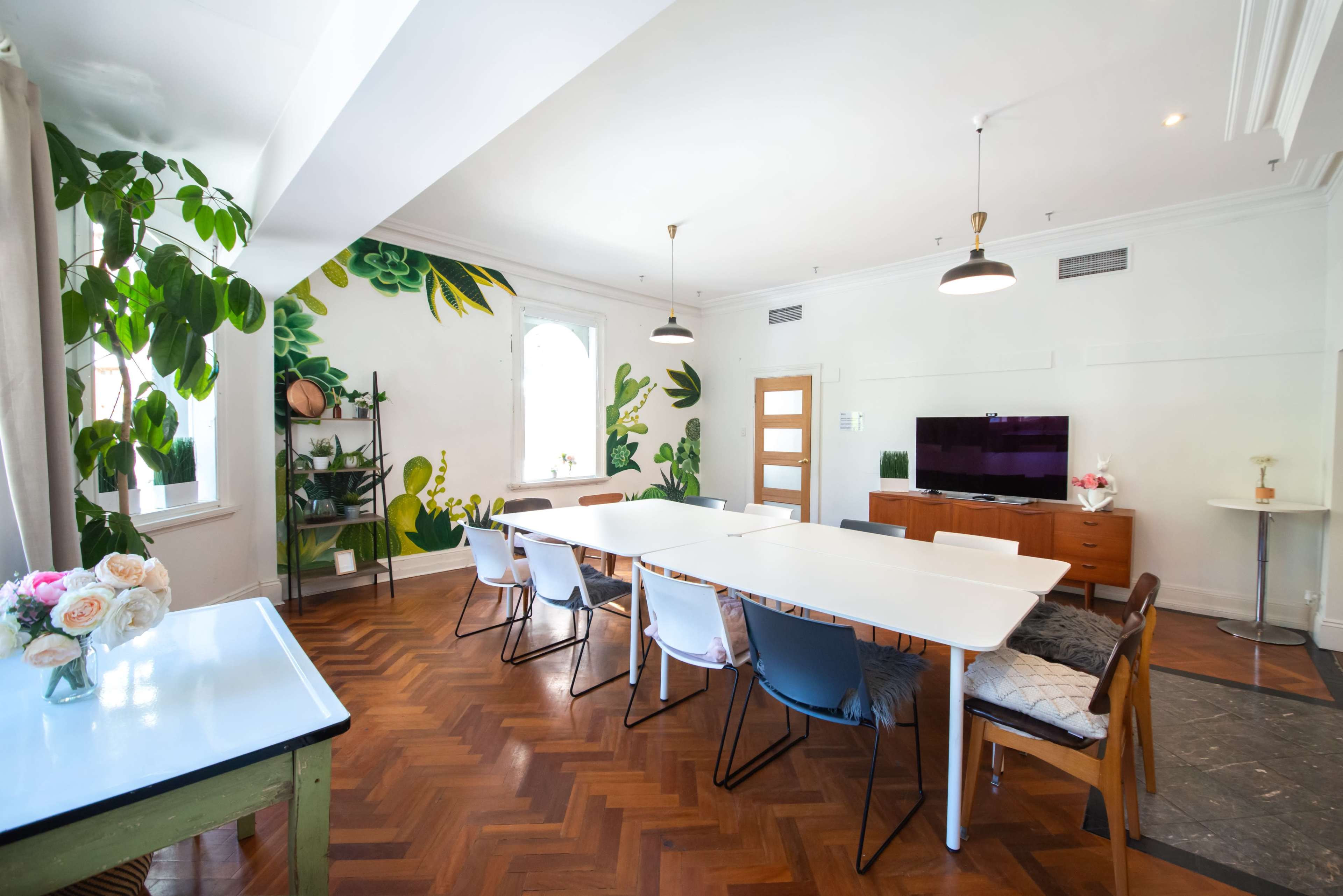 A well-lit meeting room with a long white table surrounded by chairs, a television on a stand, and a wall featuring a green leafy mural.