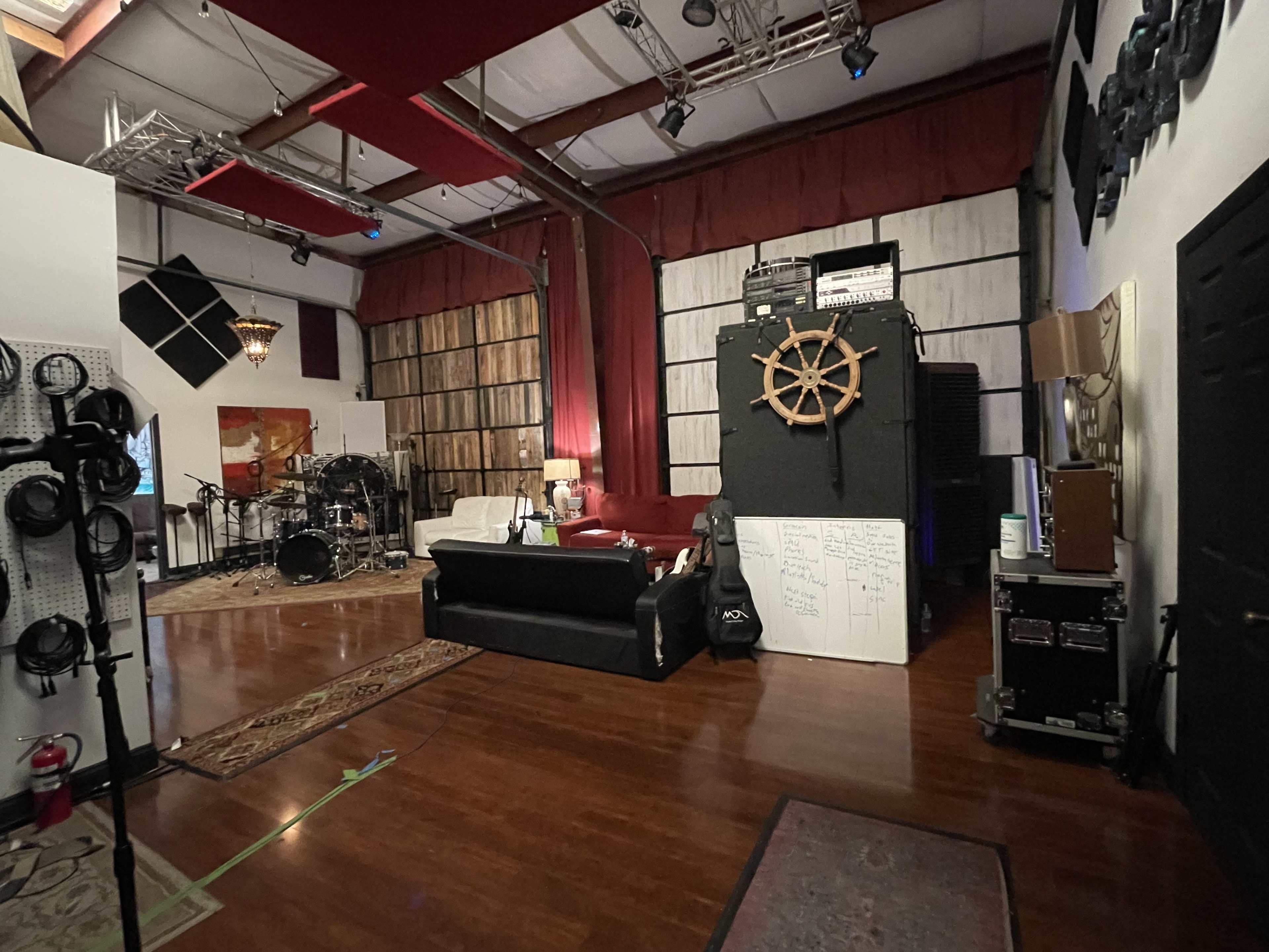 A music studio with a wooden floor, a dark couch, a drum set, and an array of sound equipment against a backdrop of wooden panels and red drapes.