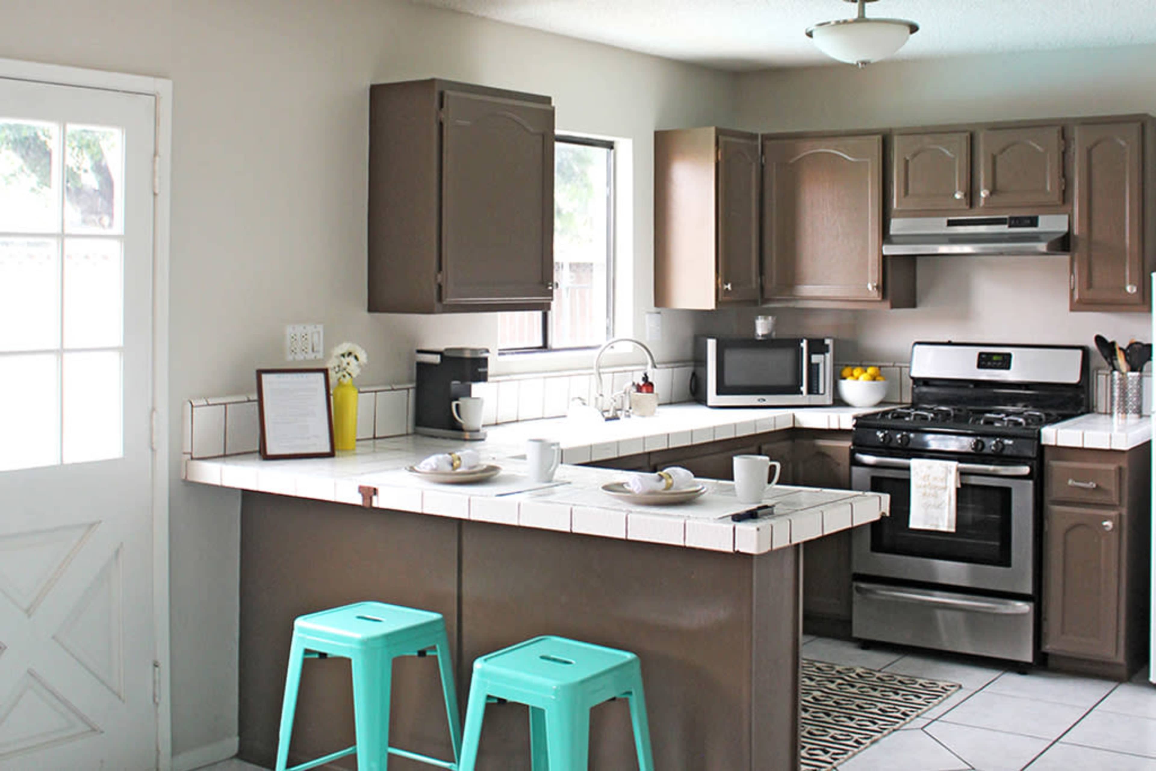 A modern kitchen features a gas stove, microwave, and two turquoise stools at a tiled island, with sunlight streaming through a window and a door leading outside.