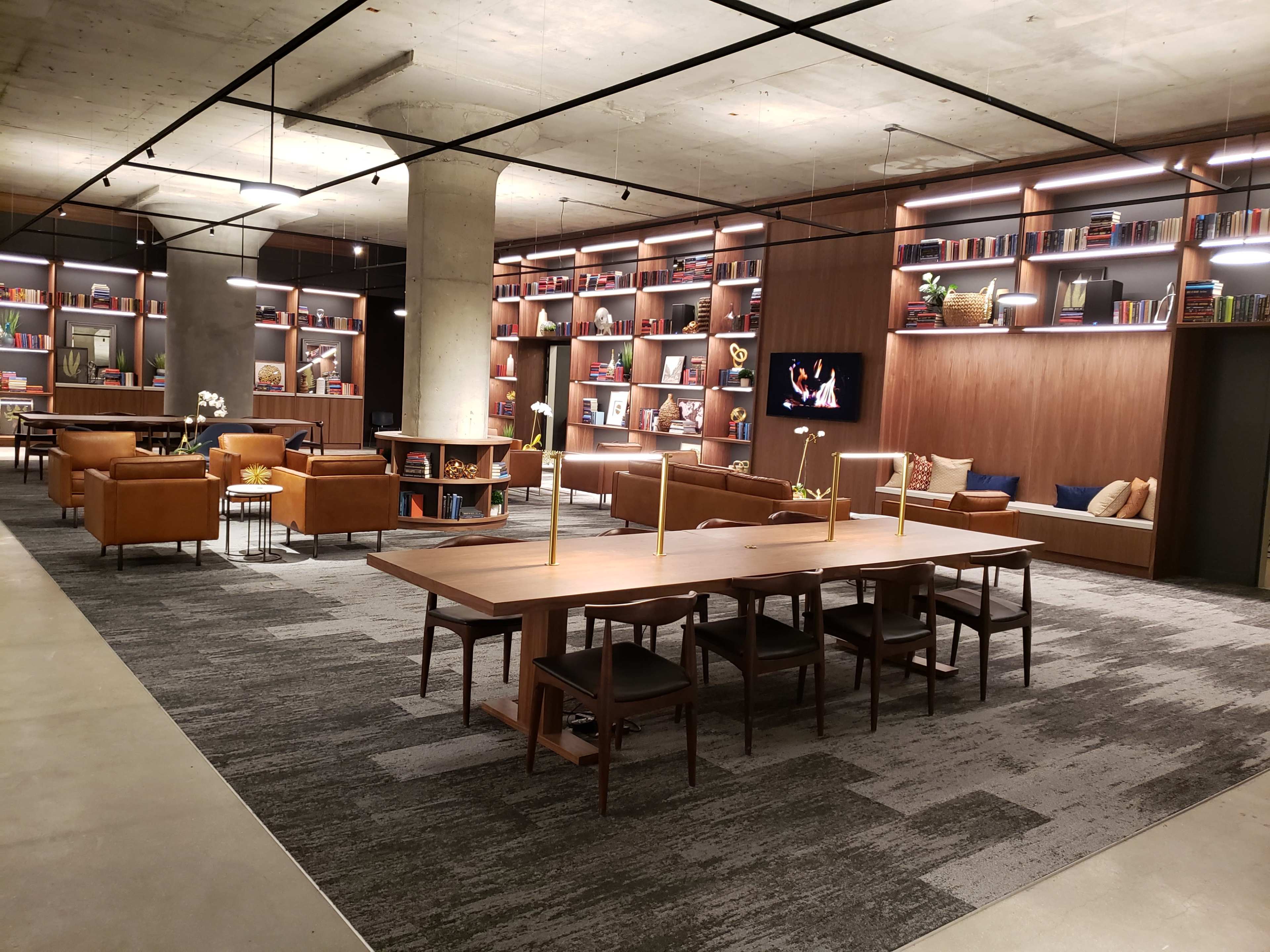 A modern library or lounge area with wooden furniture, shelves filled with books, and soft seating arranged around a central table.