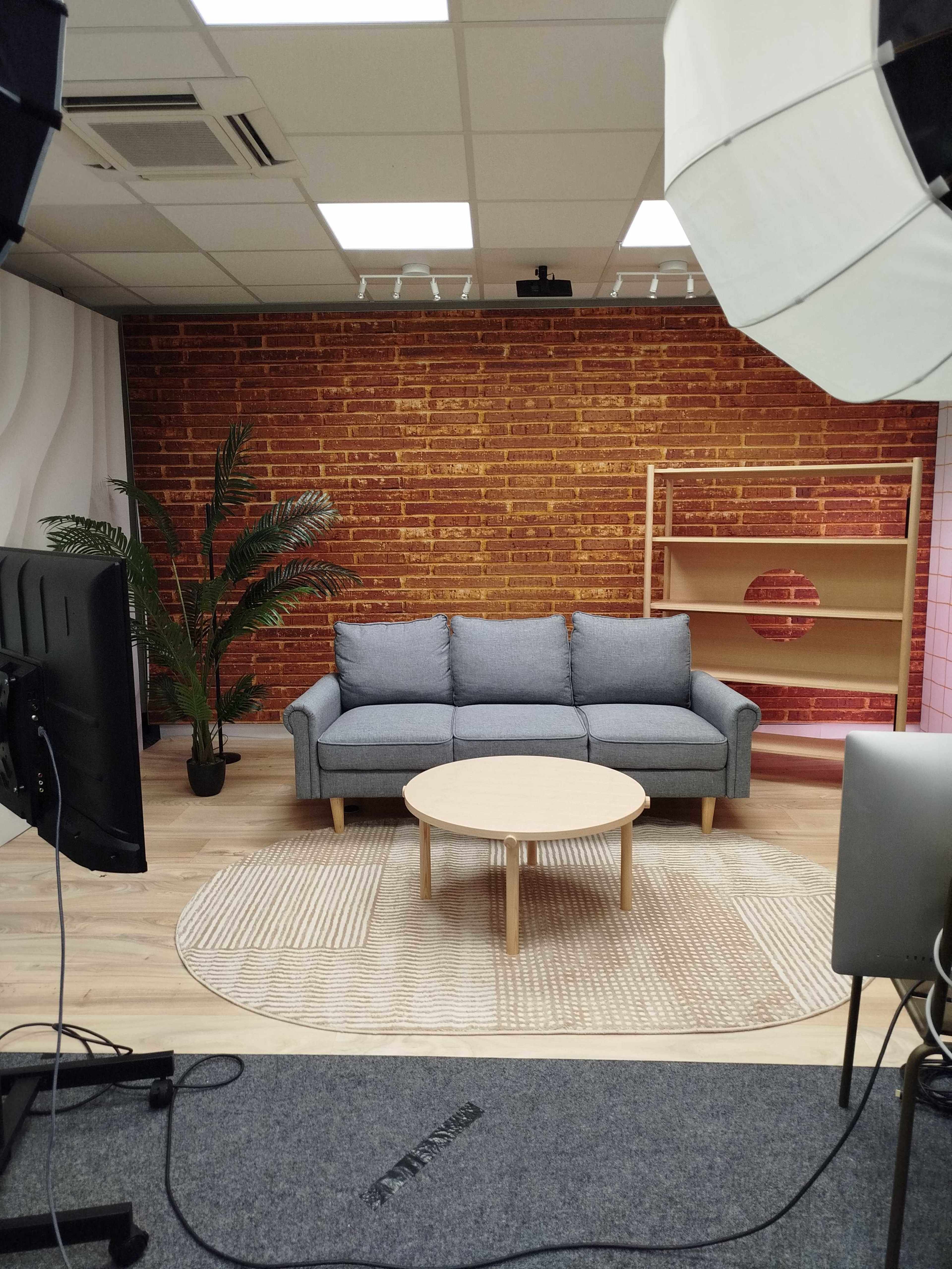 The image shows a modern studio setting with a grey sofa, a round wooden coffee table, a plant, and a brick-patterned backdrop.