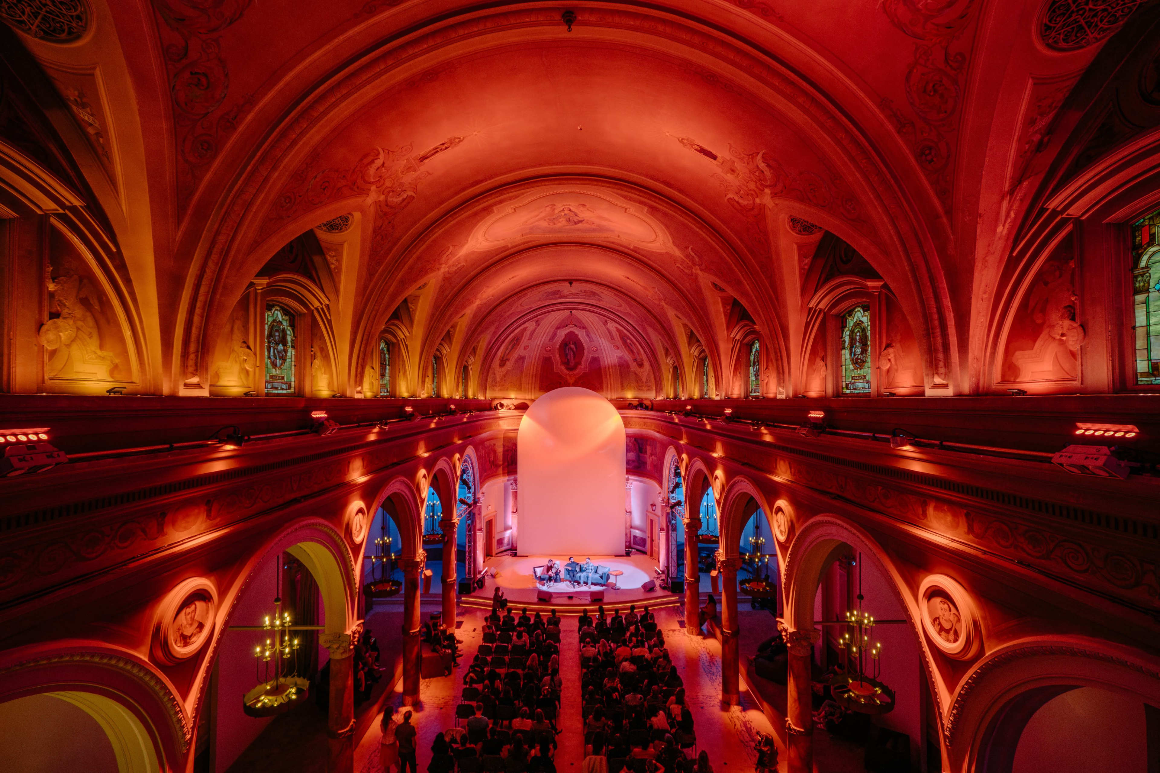 A spacious concert hall characterized by an elaborately arched ceiling and a stage set in an illuminated area with an audience seated below.