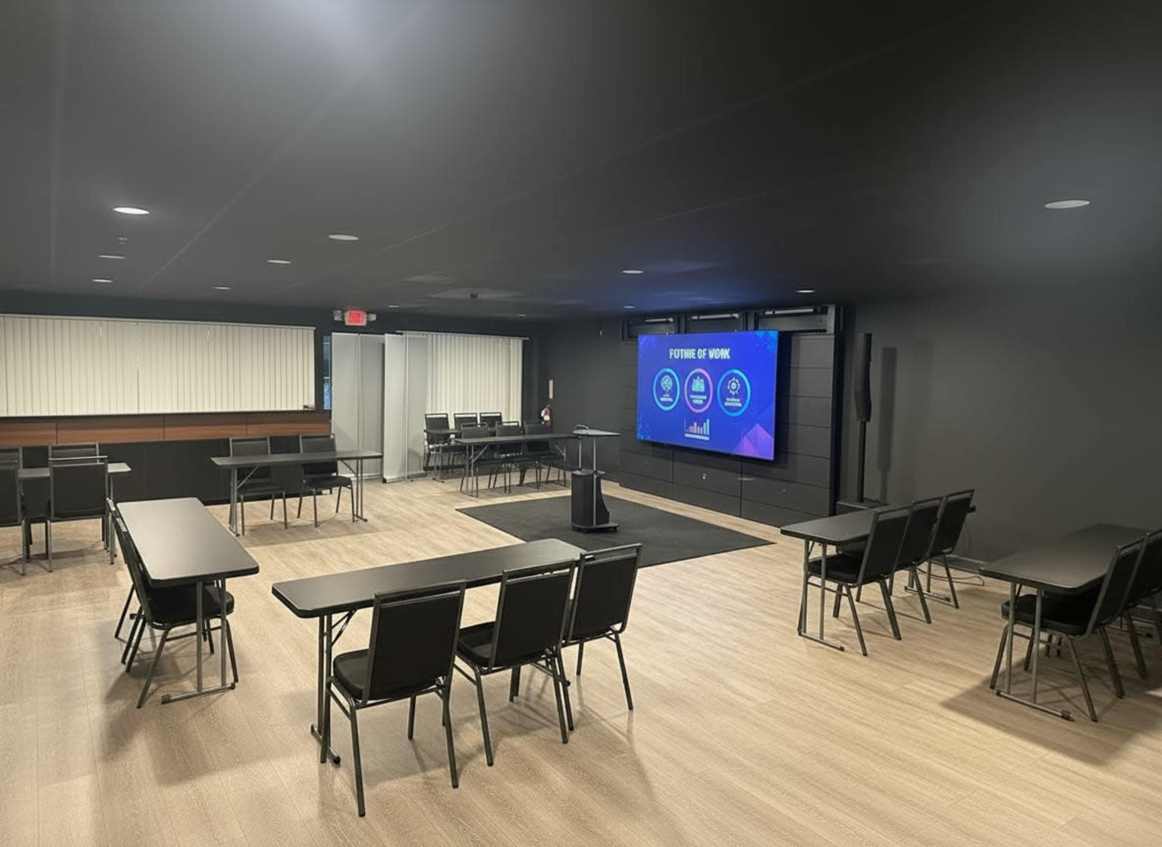 The image shows a modern conference room with multiple rows of tables and chairs, a large screen displaying a presentation, and windows with blinds.