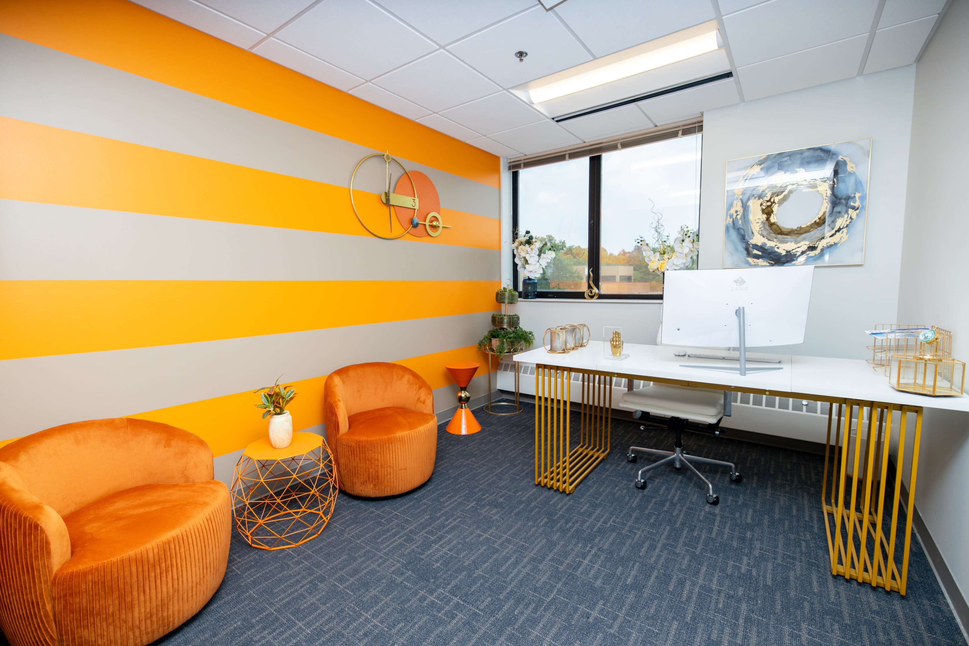 A modern office space with orange and gray striped walls, two orange chairs, a white desk with a monitor, and decorative plants.