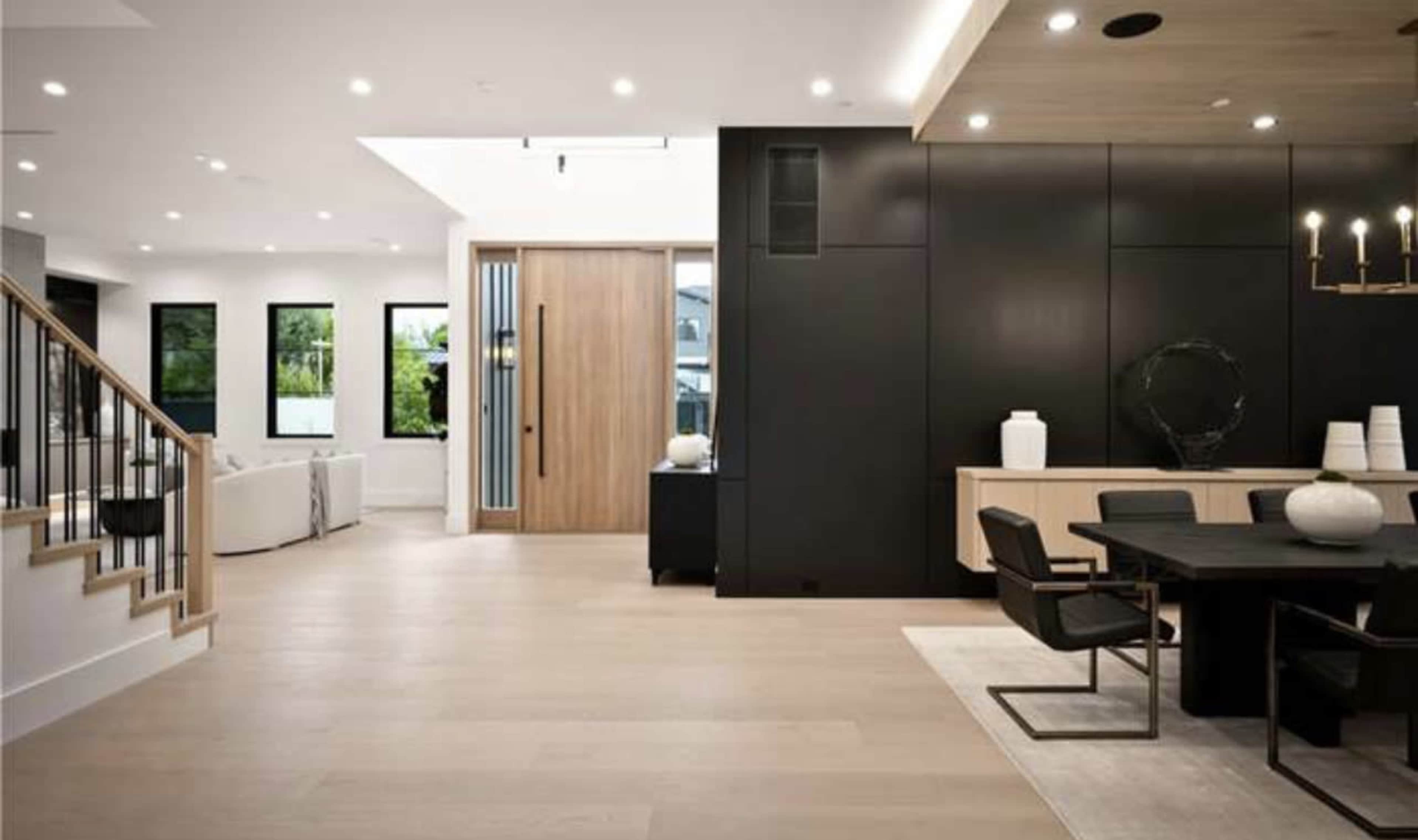 A modern interior space featuring an open floor plan with a wooden front door, a dining area with a black table and chairs, and a staircase leading to the upper level.
