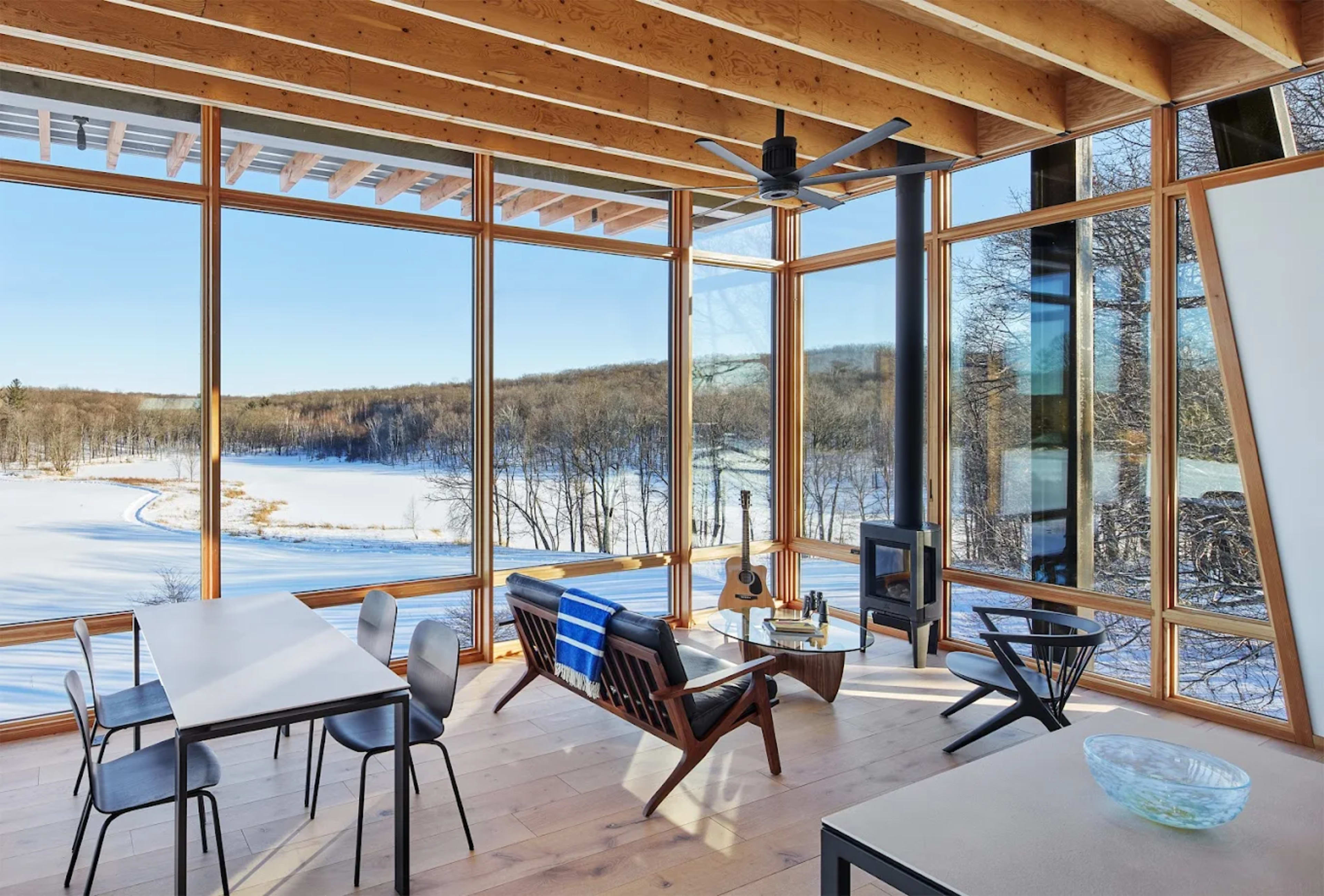 The image features a modern, glass-walled living space with panoramic views of a snowy landscape, furnished with a table, chairs, and a sectional sofa near a fireplace.