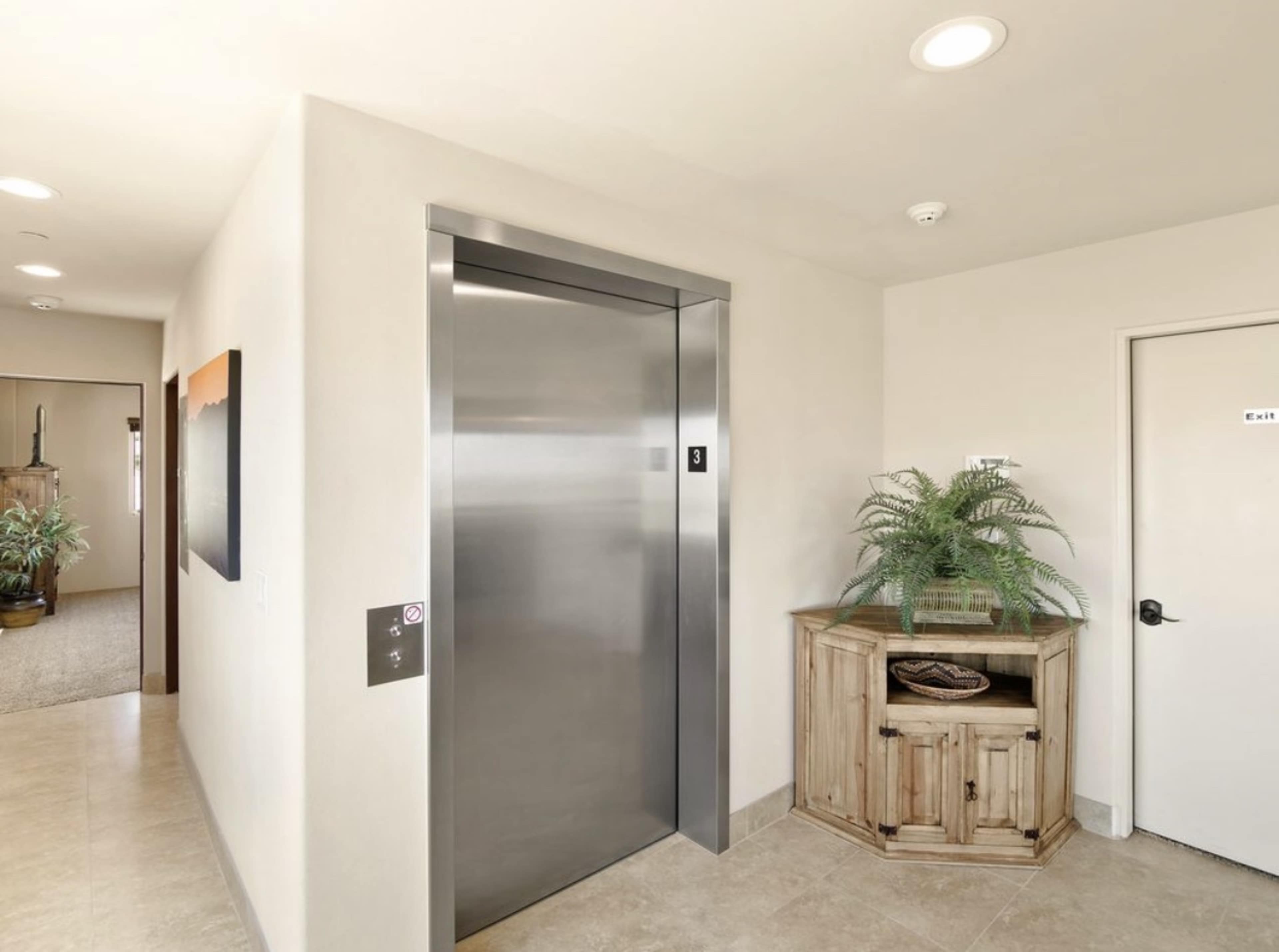 A modern elevator entrance in a well-lit hallway alongside a wooden cabinet and a potted plant.