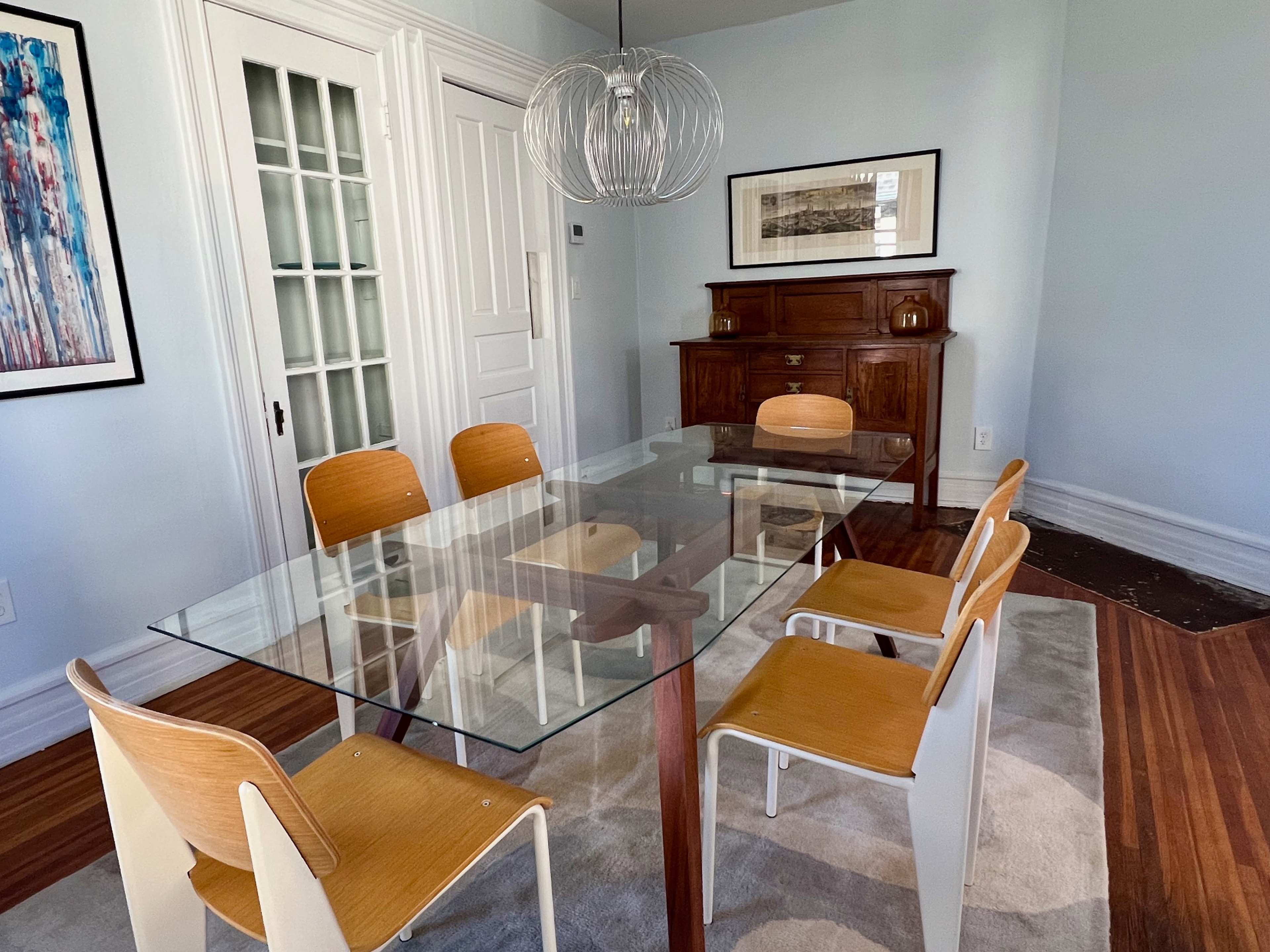A modern dining room features a glass-topped table surrounded by four wooden chairs and a wooden cabinet against the wall.