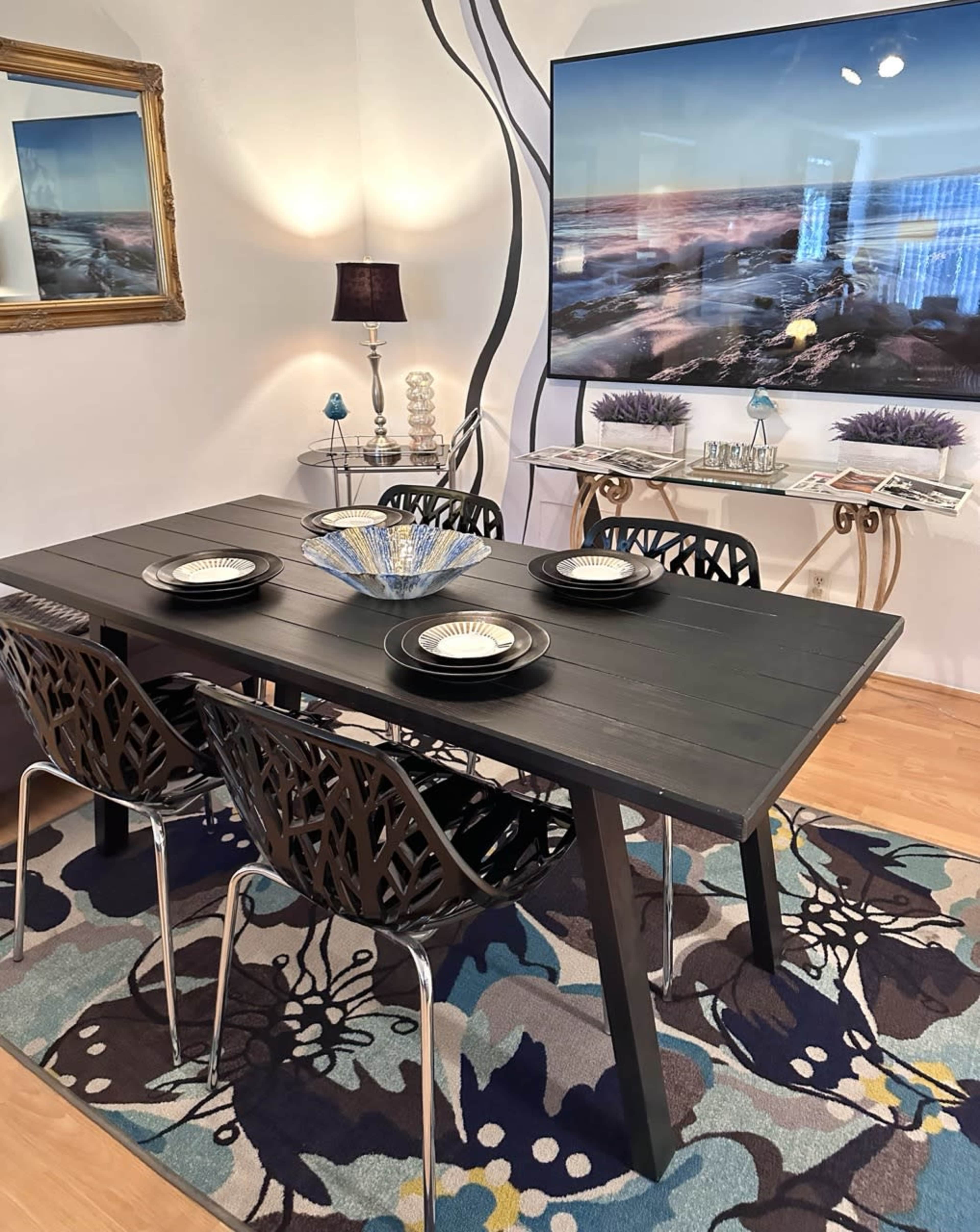 A dining room scene features a black table set with plates and surrounded by modern chairs, with a large wall-mounted screen displaying a landscape and decorative items on an adjacent table.