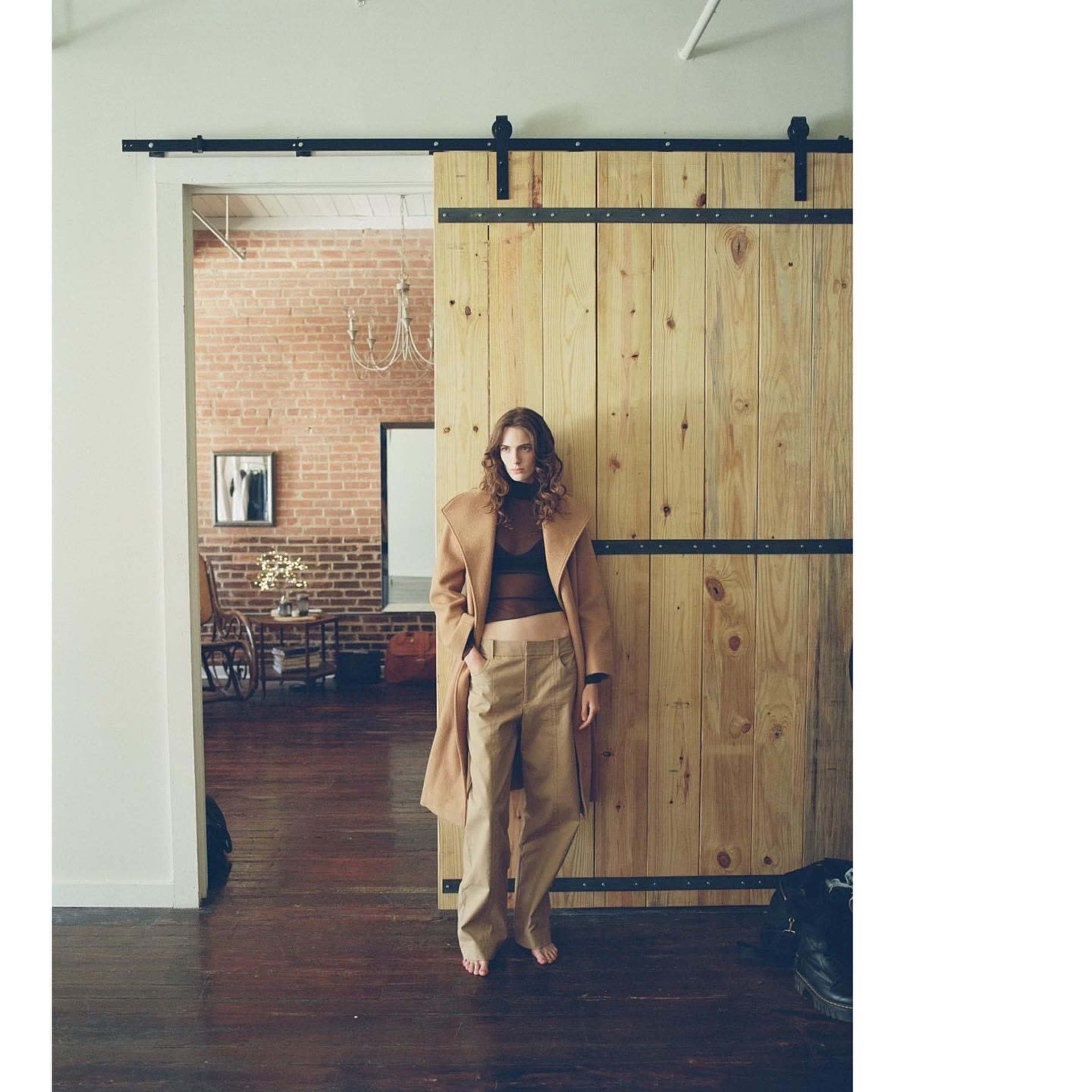 A model stands barefoot in front of a large wooden sliding door in a room with exposed brick walls and a vintage chair.