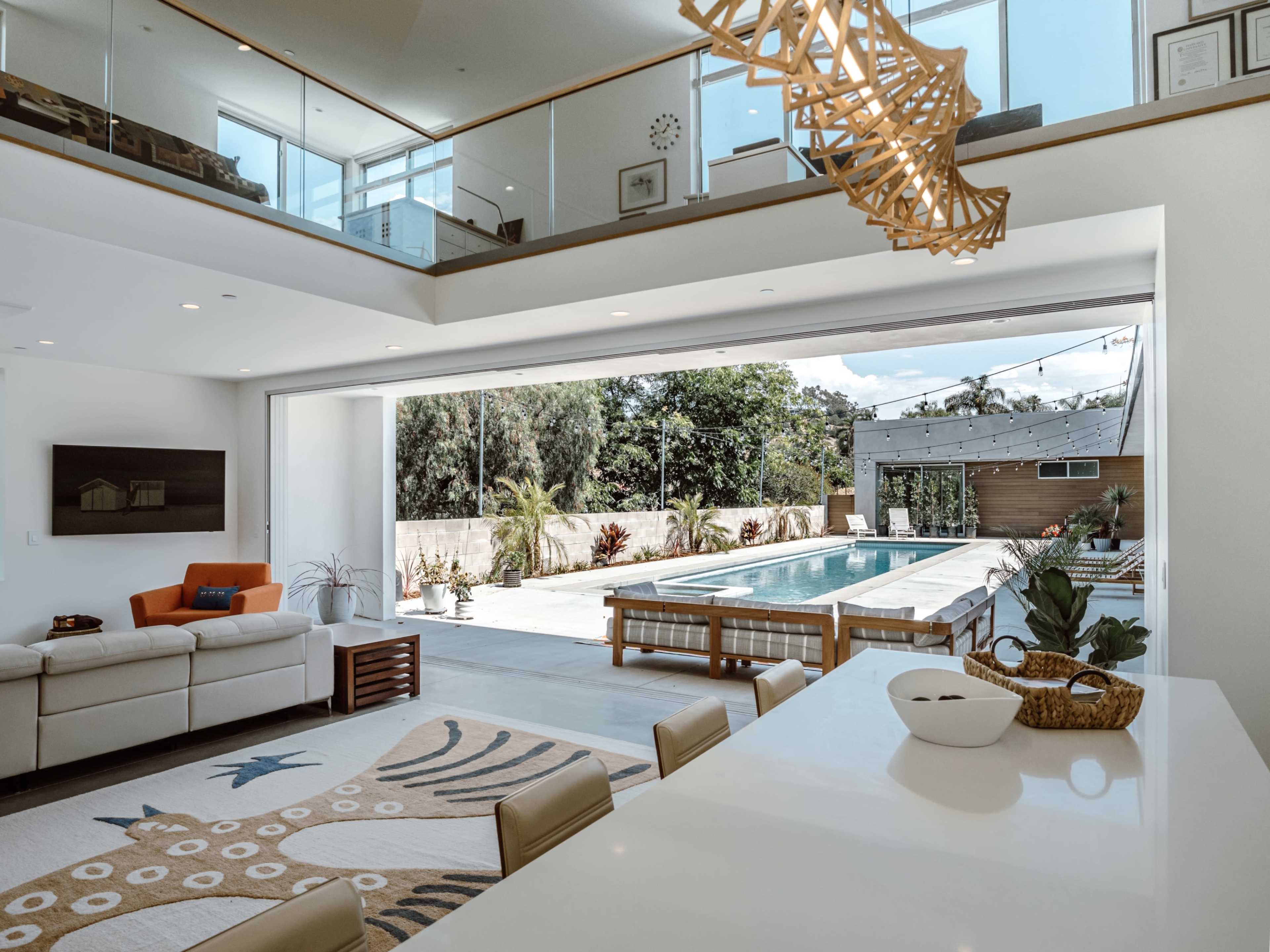 The image shows a modern open-concept living area leading to an outdoor pool, with large sliding glass doors and minimalistic furnishings.