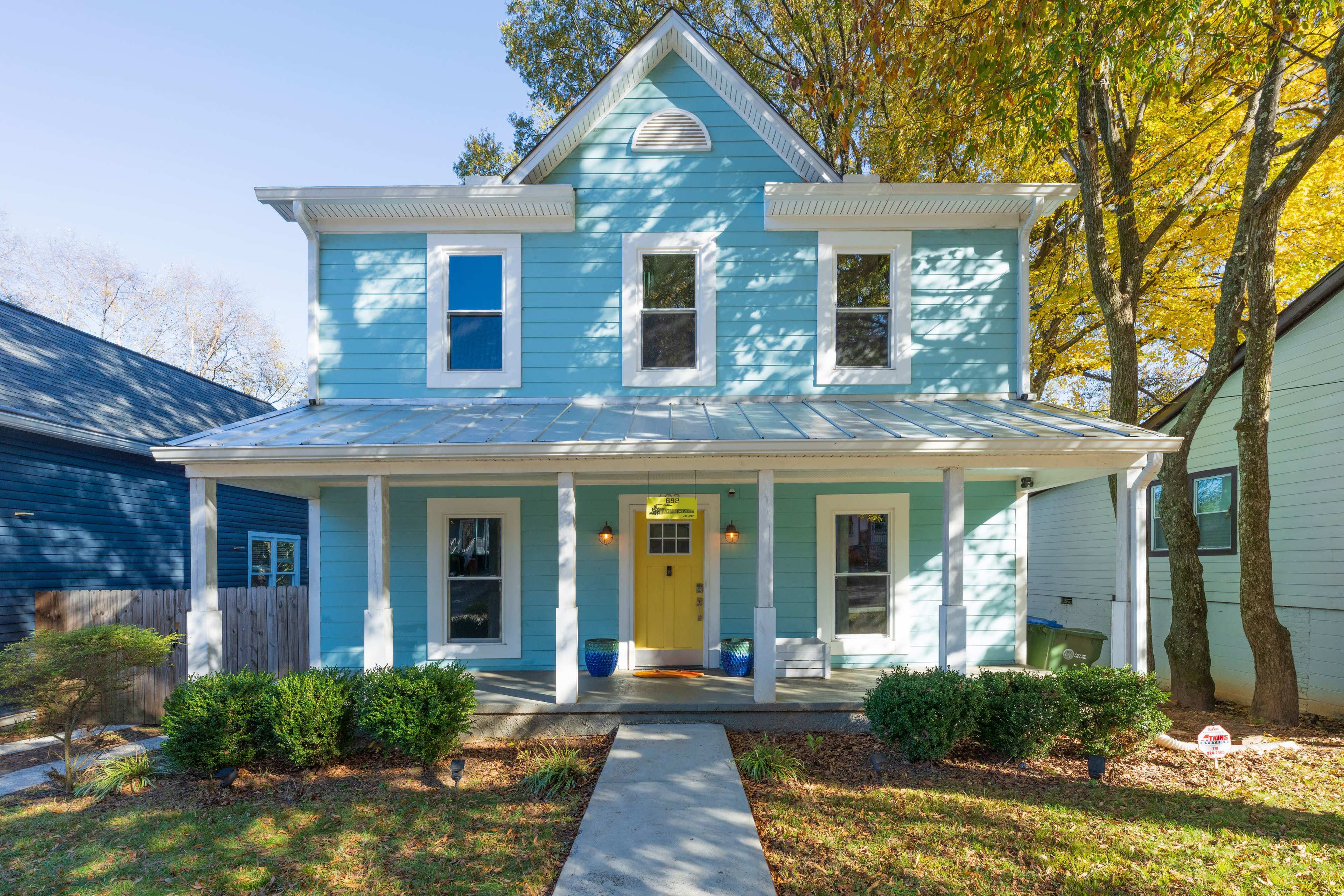 A two-story blue house with white trim features a front porch, green shrubs, and a well-maintained pathway.