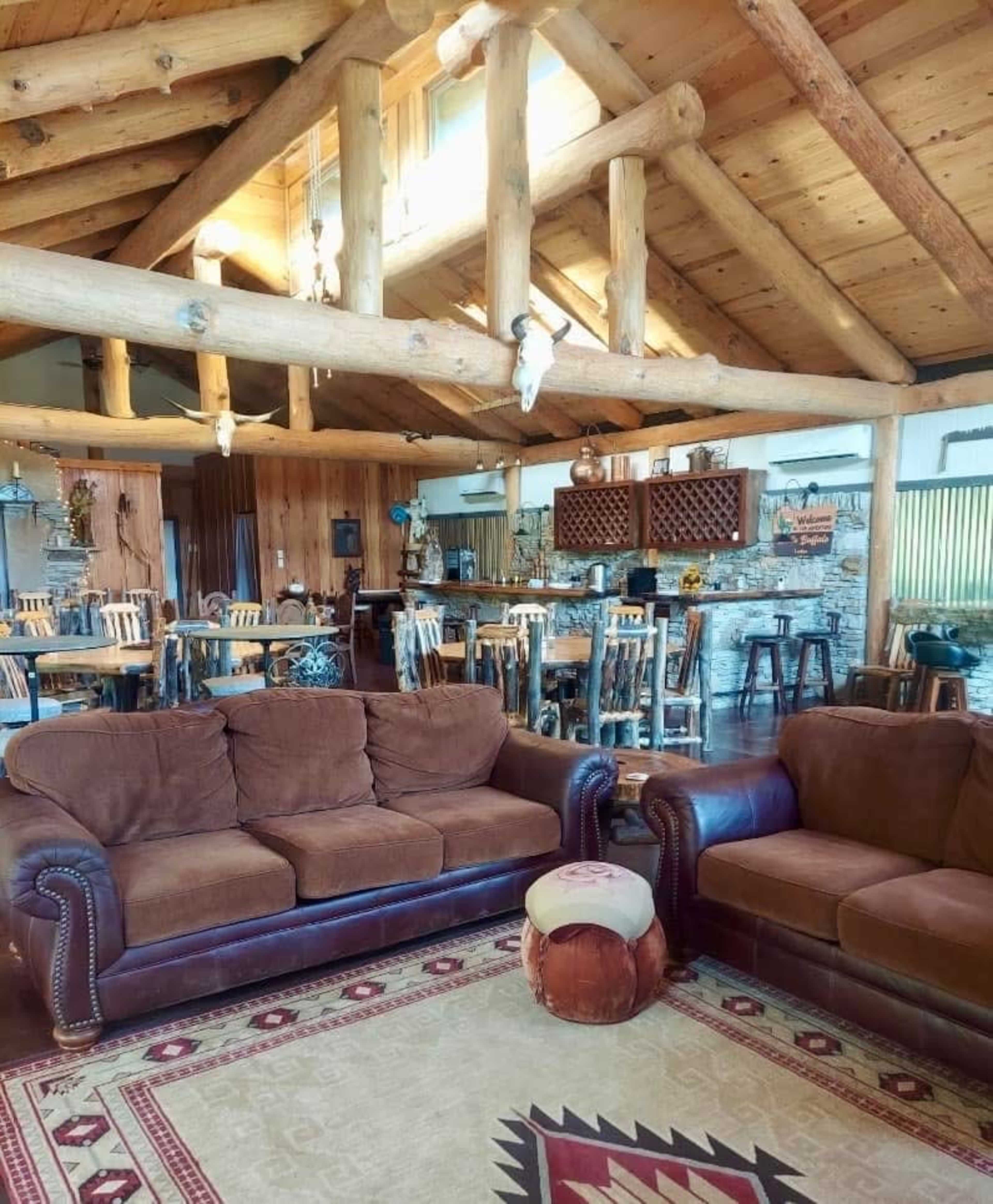 The image shows a spacious, rustic living area featuring wooden beams, a cozy seating arrangement with brown sofas, and a dining area in the background.