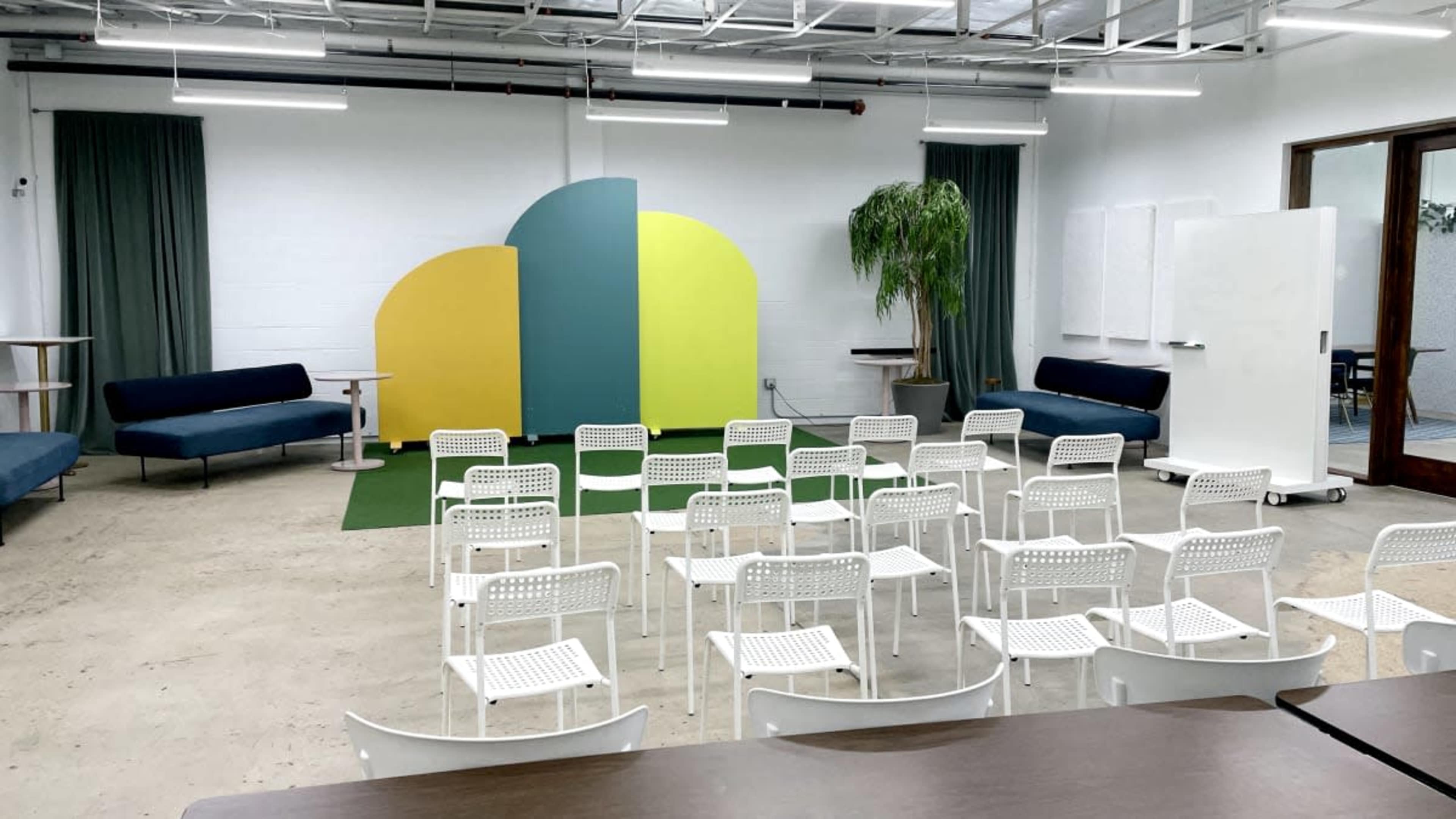 The image shows a modern event space with white chairs set up in rows facing a colorful backdrop, accompanied by seating areas and plants.